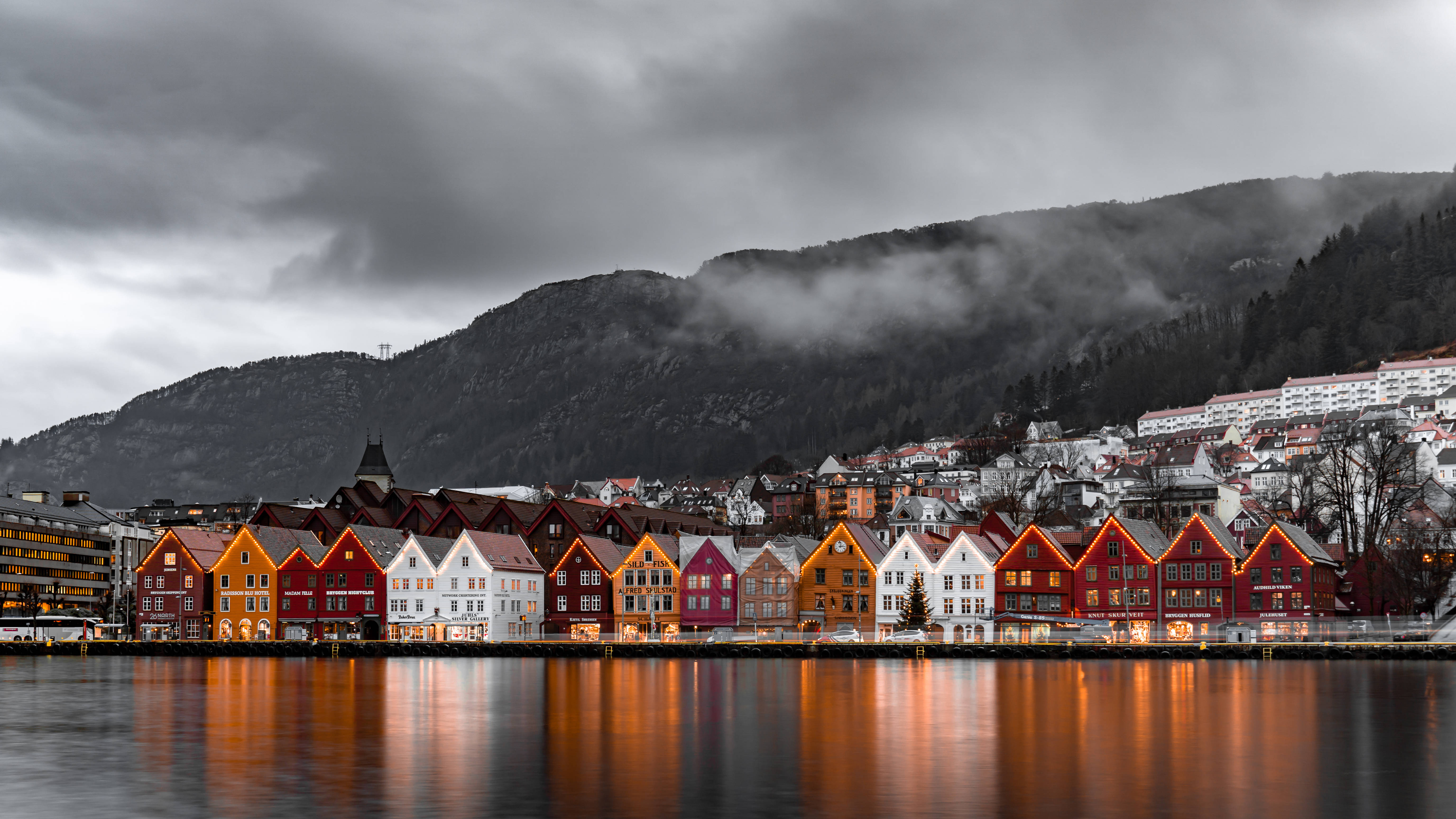 Norway 桌布 壁紙 — 鮮豔的紅色、橙色和白色木屋排列在平靜的海濱，在陰沉的灰色天空下反射著溫暖的燈光，背後是霧濛濛的山脈。