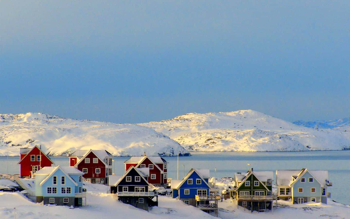Greenland 桌布 壁紙 — 鮮豔的紅色、藍色和綠色房屋點綴在雪覆蓋的海濱，遠處是白雪覆蓋的山脈，天空清澈湛藍。