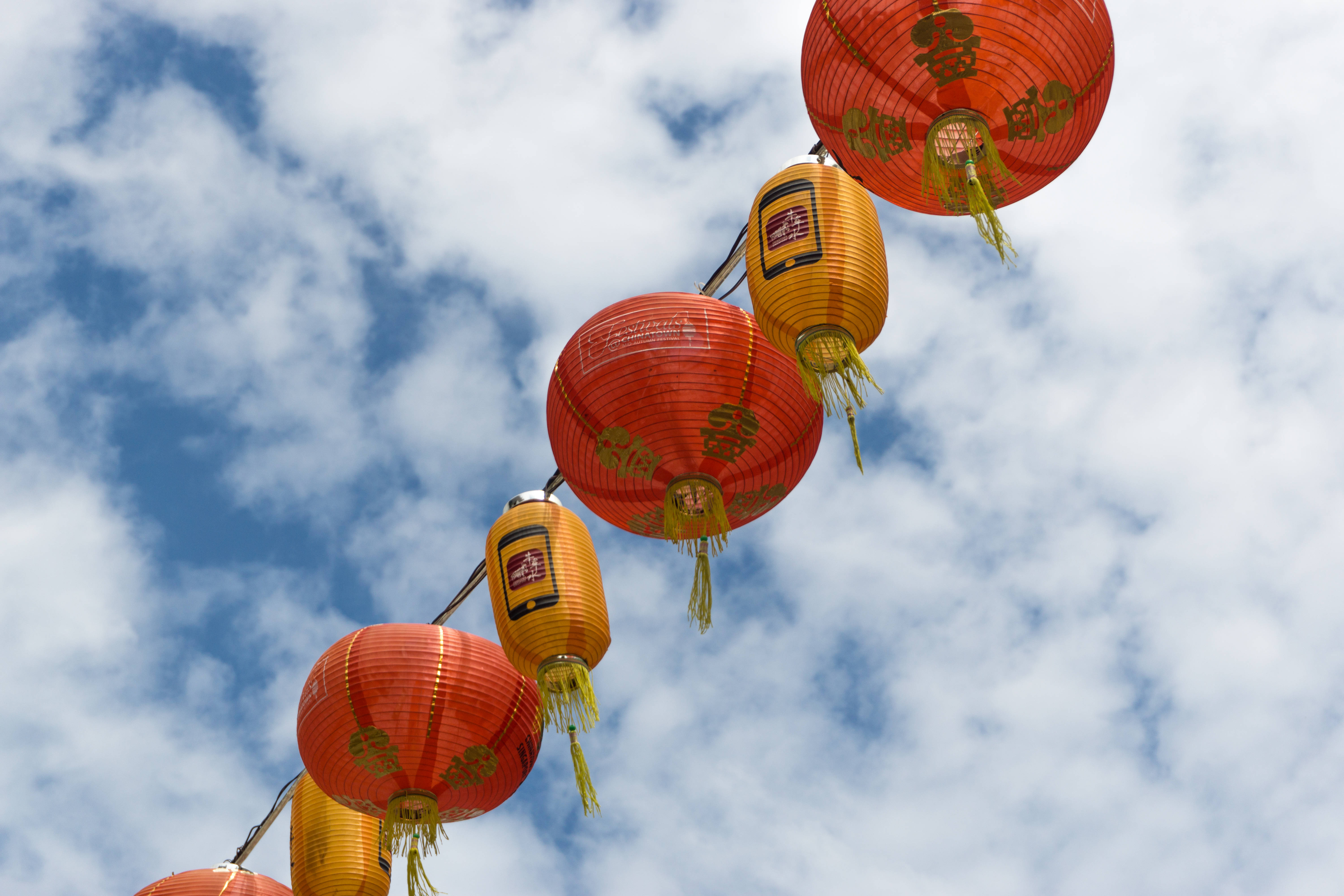 春節 桌布 壁紙 — 鮮豔的紅色和橙色紙燈籠帶有金色流蘇，掛在點綴著蓬鬆白雲的明亮藍天上。