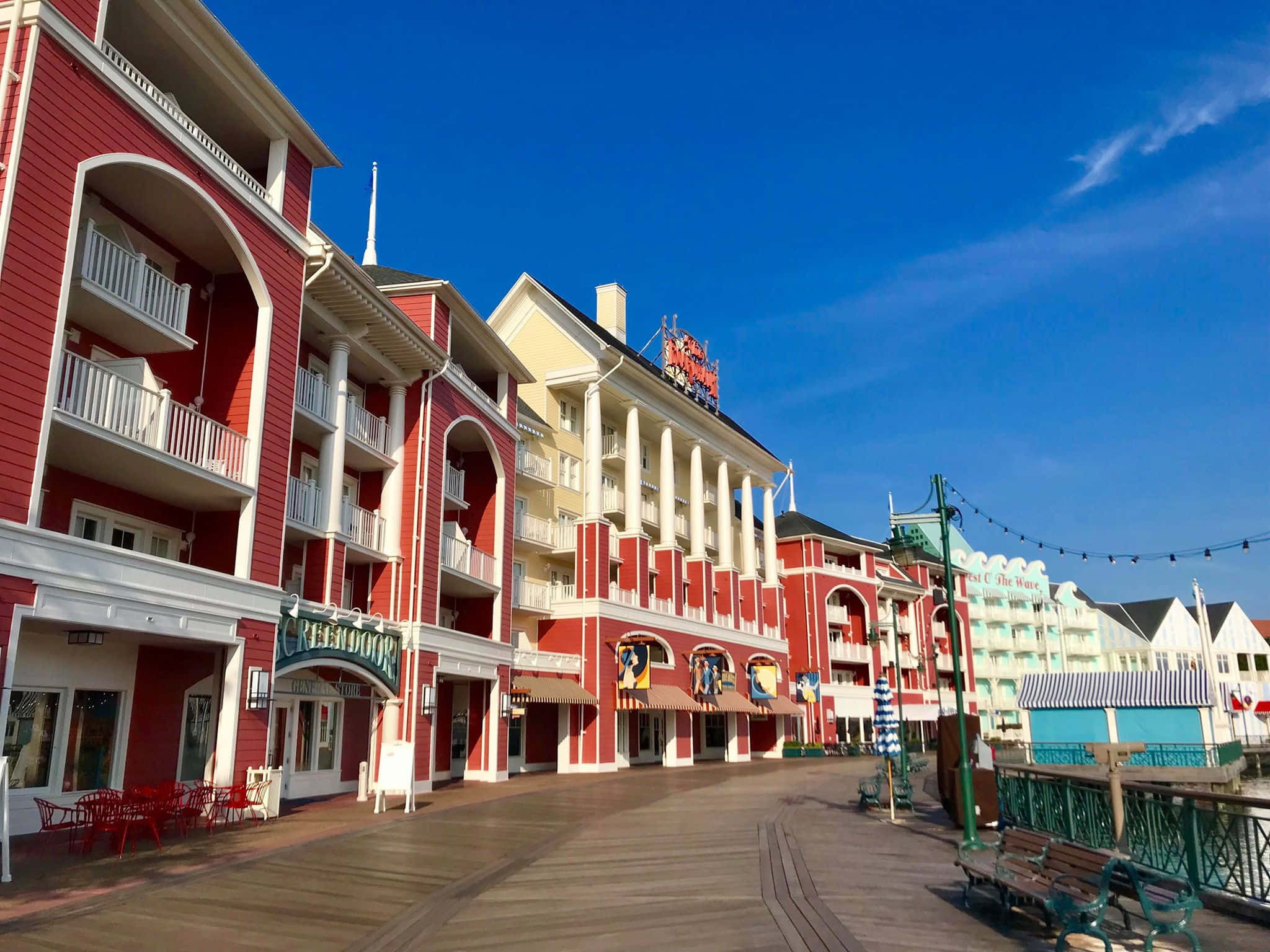 Boardwalk 桌布 壁紙 — 鮮豔的紅色和白色度假村建築沿著木質木板路排列，天空湛藍清澈，白雲點點。