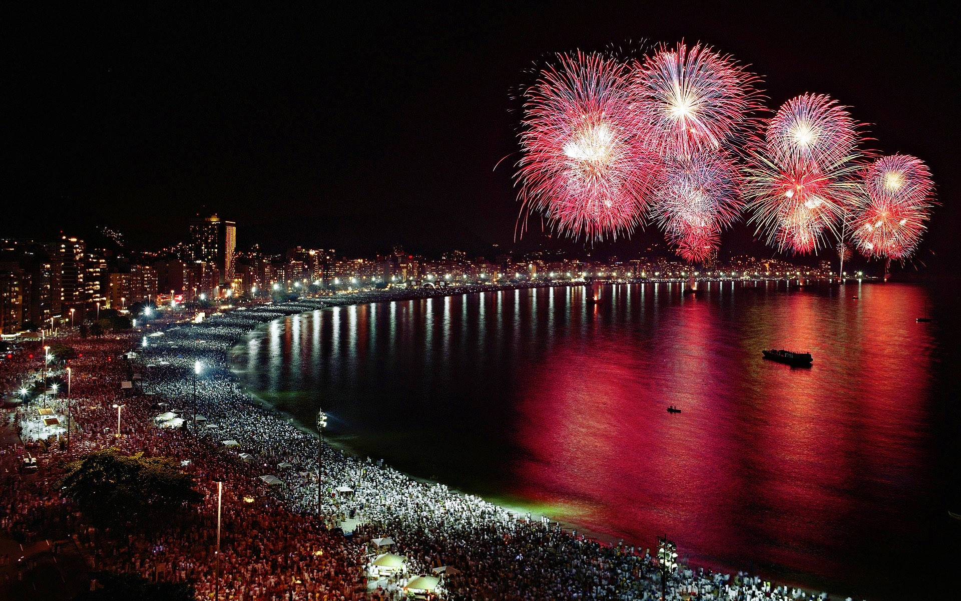 Fireworks 桌布 壁紙 — 鮮豔的紅色和白色煙火在夜間繁華的海岸城市景觀上空爆炸，照亮了下面的水域和人群。