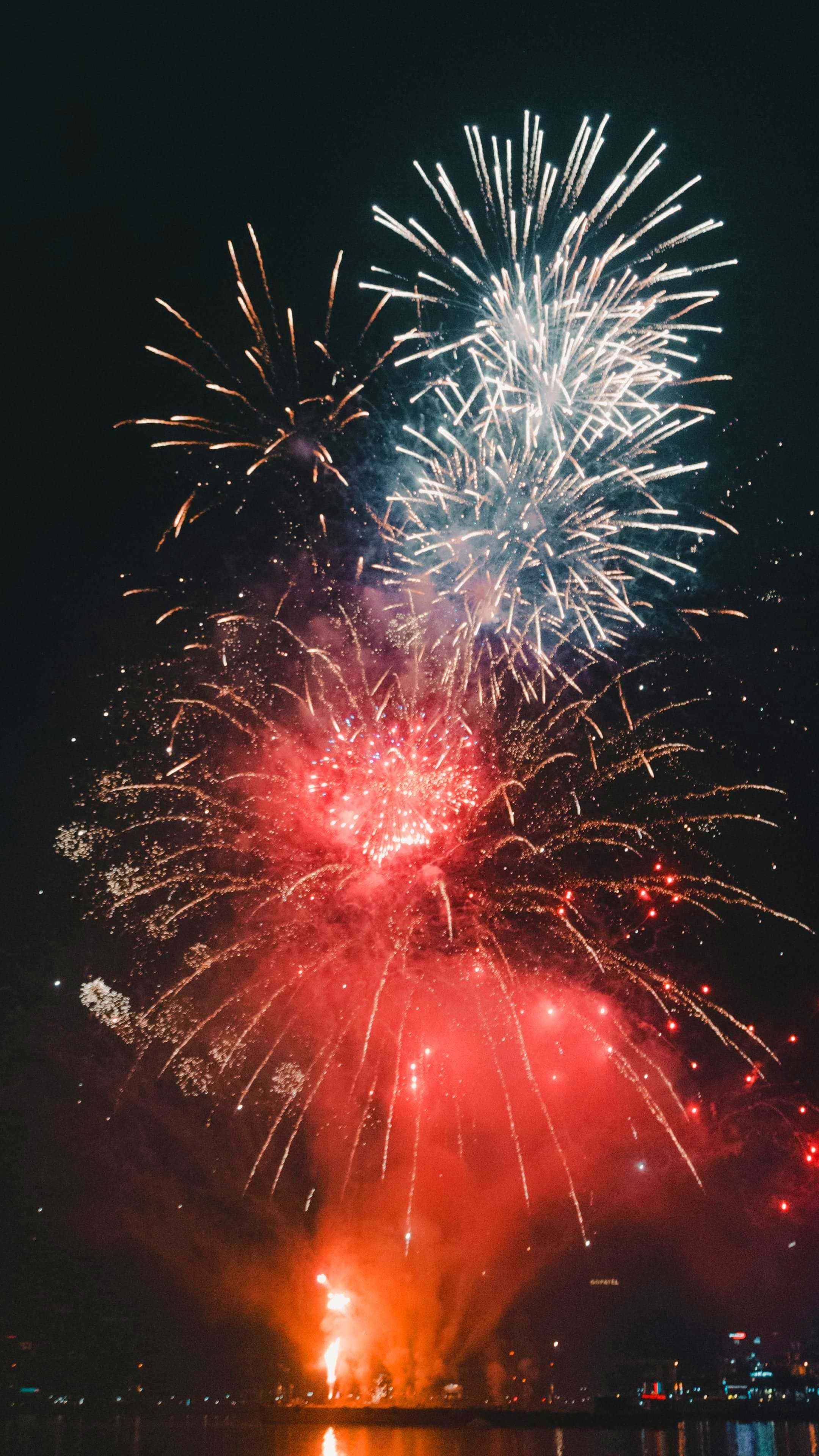 Fireworks 桌布 壁紙 — 鮮豔的紅色和白色煙火在漆黑的夜晚水面上爆炸，倒映著城市的燈光。