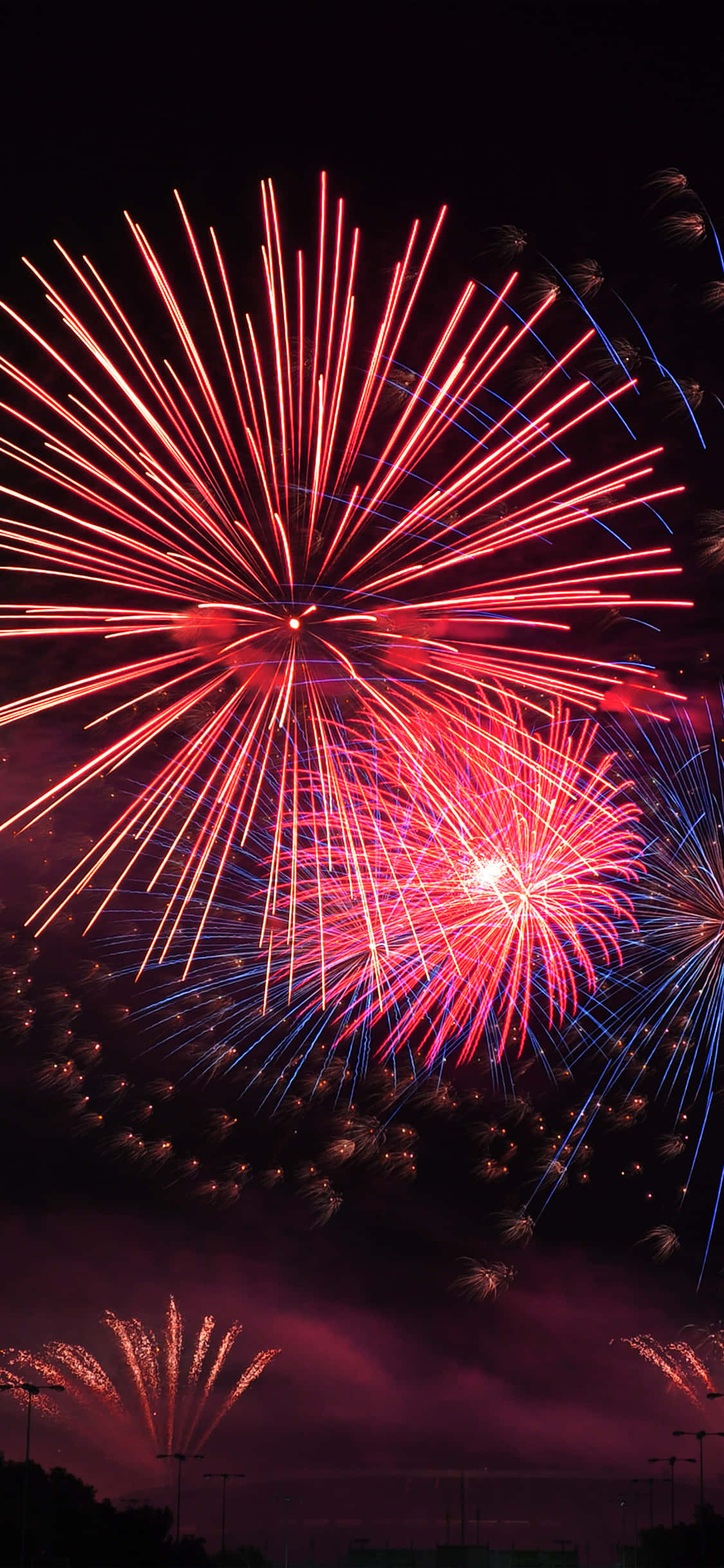 firework 桌布 壁紙 — 鮮豔的紅色和藍色煙火在遙遠城市景觀上方的黑暗夜空中綻放出耀眼的光芒。