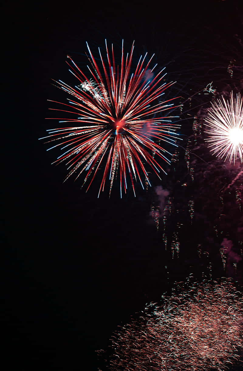 Happy New Year Phone 桌布 壁紙 — 鮮豔的紅色和藍色煙火在黑暗的夜空中爆炸，閃爍的水面反射出它們燦爛的光芒。