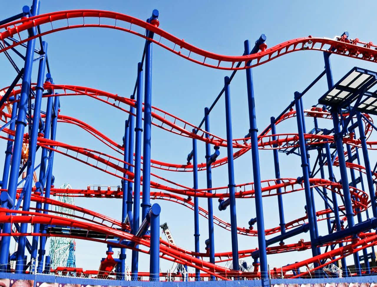 Roller Coaster 桌布 壁紙 — 鮮豔的紅色和藍色雲霄飛車在遊樂園的清澈藍天下蜿蜒盤旋並翻滾。