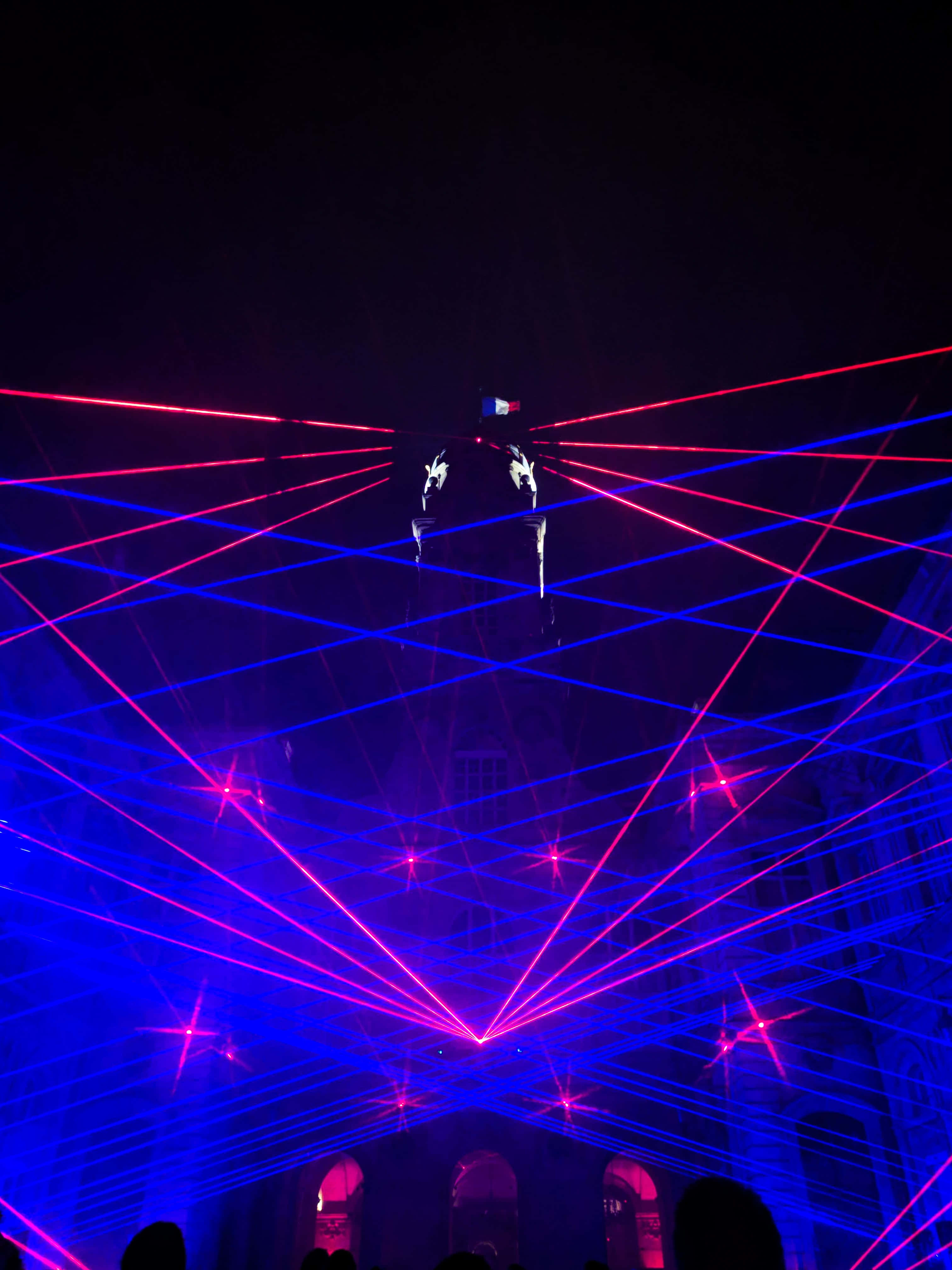 Laser 桌布 壁紙 — 鮮豔的紅色和藍色雷射光束在黑暗場地的天花板上交錯，懸在剪影般的觀眾頭頂上方。