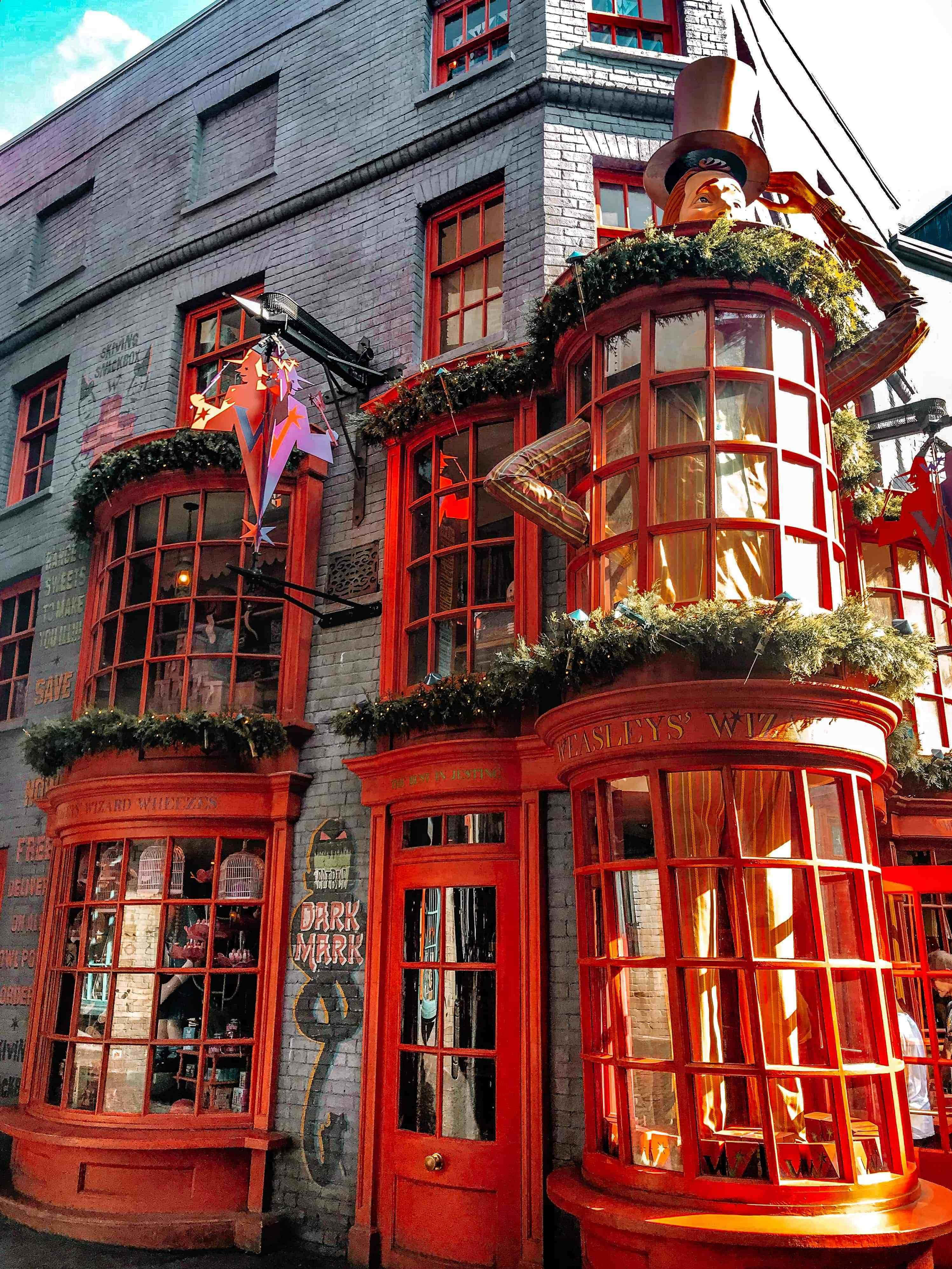 Diagon Alley 桌布 壁紙 — 鮮豔的紅色巫師商店門面，在明亮的藍天下裝飾著節日花環和奇特的裝飾。