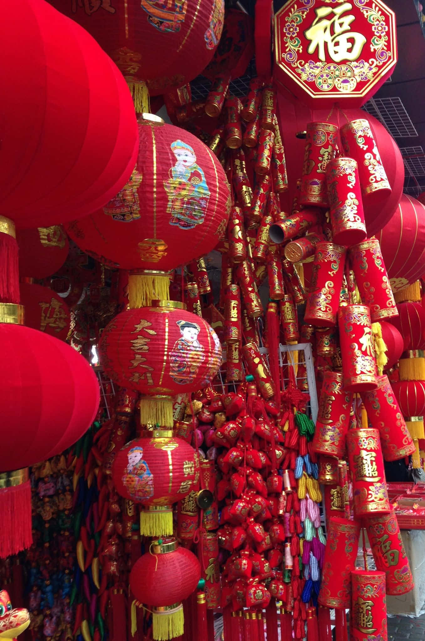 春節 桌布 壁紙 — 鮮豔的紅色燈籠和鞭炮，飾有金色中文字和傳統圖案，密密麻麻地掛在節日市場攤位上。