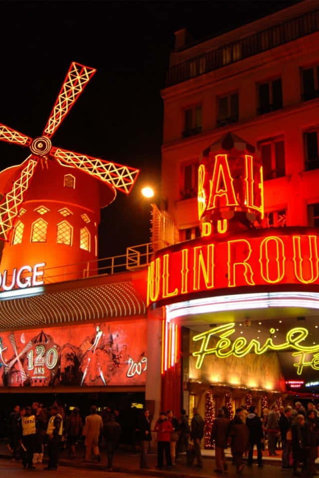 moulin rouge 桌布 壁紙 — 鮮豔的紅色霓虹燈在夜晚照亮紅磨坊，閃爍的風車和下方聚集的人群。