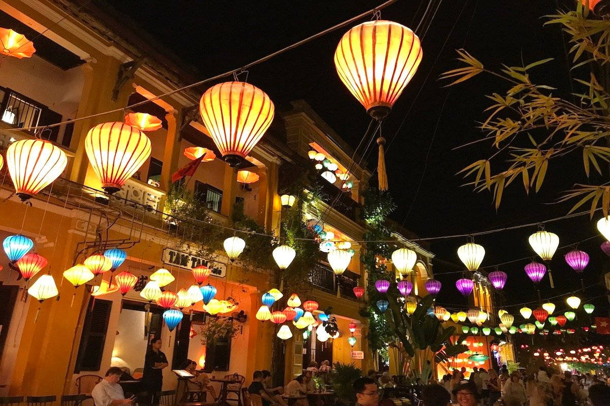 Hanoi 桌布 壁紙 — 鮮豔的紙燈籠在黑暗的夜空下溫暖地發光，照亮了一條兩旁種滿黃色建築物和竹樹的繁華街道。