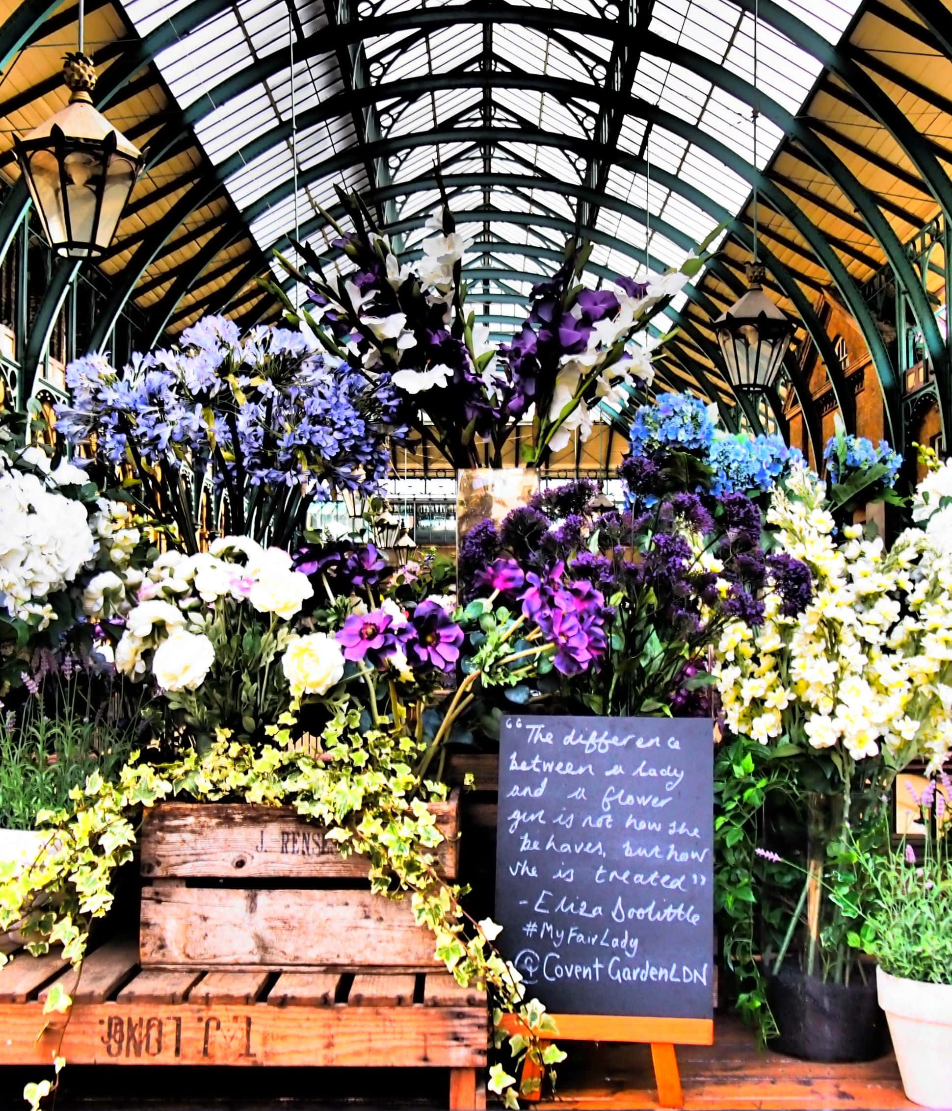 Flower Market 桌布 壁紙 — 鮮豔的紫色、白色和黃色花朵在玻璃屋頂市場大廳的木箱中盛開，掛著燈籠。