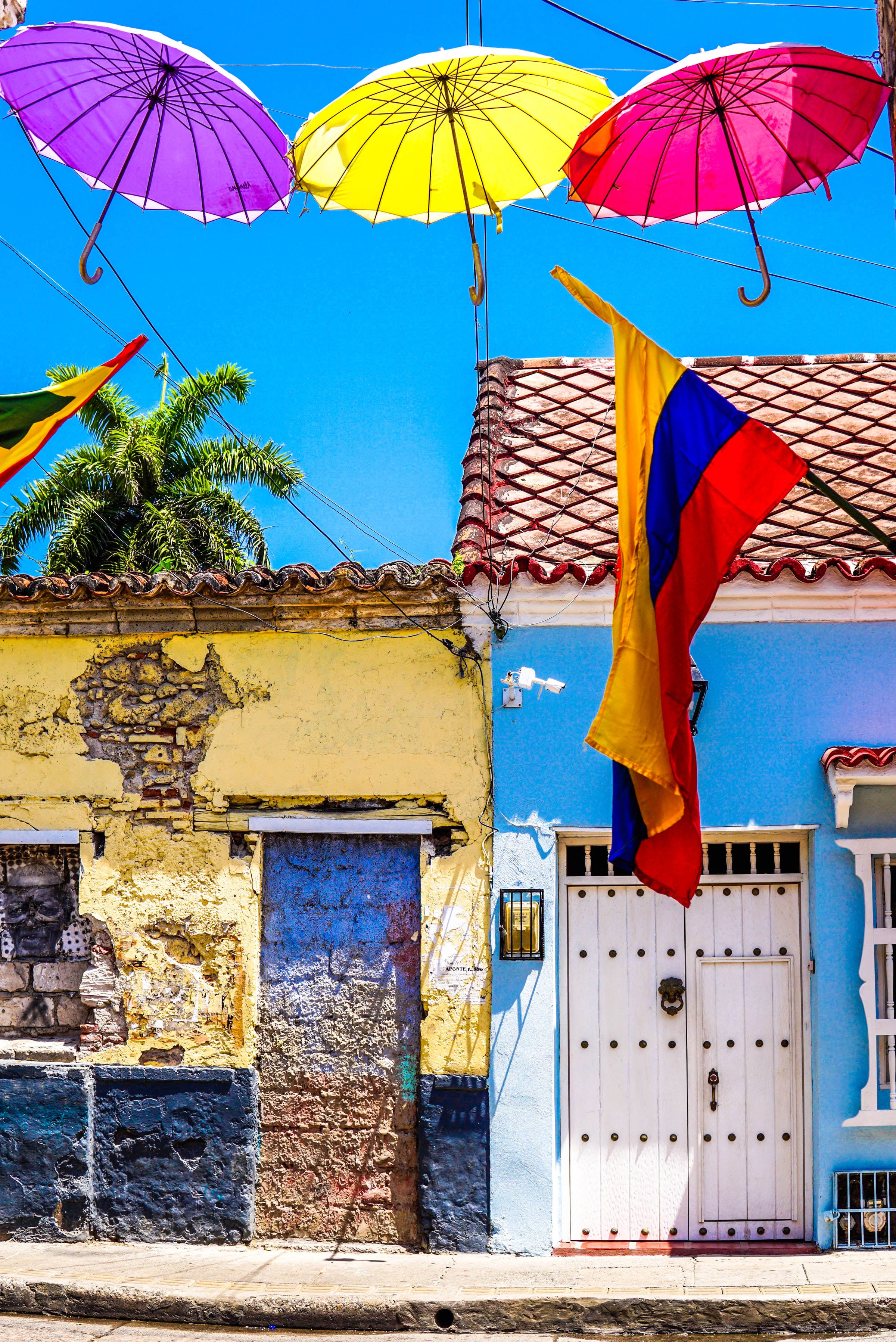 Colombia Flag 桌布 壁紙 — 鮮豔的紫色、黃色和粉紅色雨傘懸掛在色彩繽紛的殖民建築上方，在晴朗的藍天下，哥倫比亞國旗飄揚。