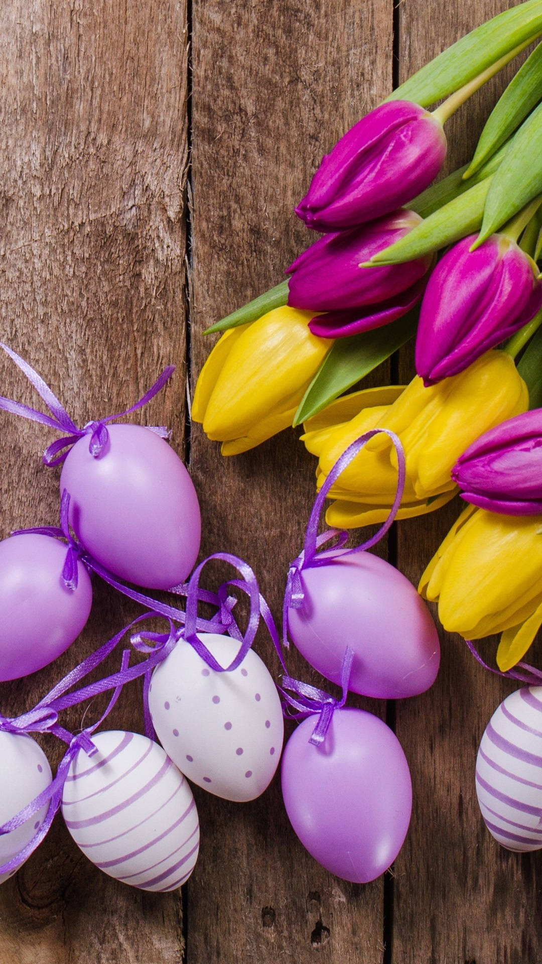 Easter iPhone 桌布 壁紙 — 鮮豔的紫色和黃色鬱金香，旁邊是粉紫色、白色和條紋的復活節彩蛋，用絲帶綁著，放在質樸的木質表面上。