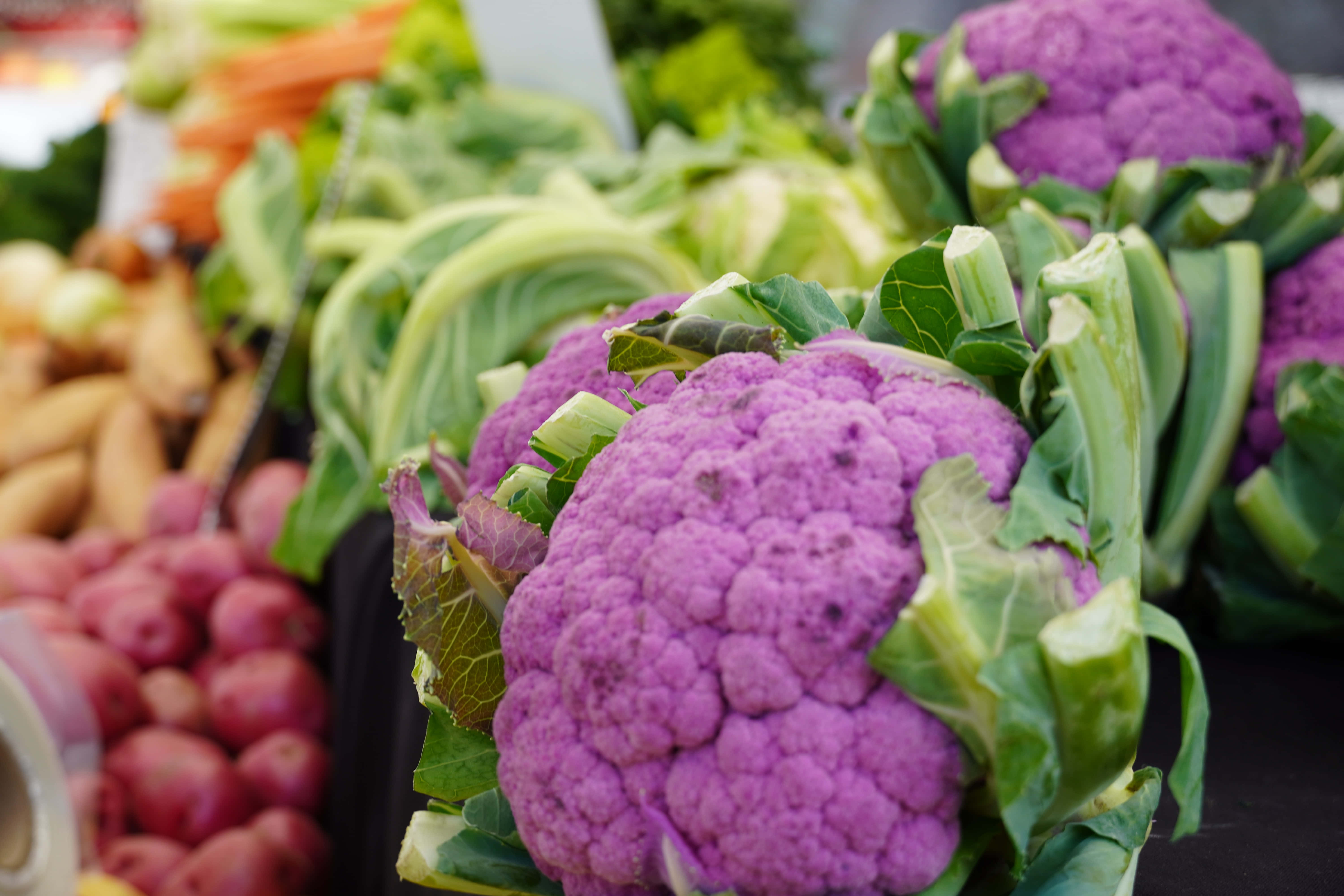 Purple Cauliflower 桌布 壁紙 — 鮮豔的紫色花椰菜搭配綠色葉子，在市場攤位上的其他色彩繽紛的新鮮蔬菜中格外顯眼。