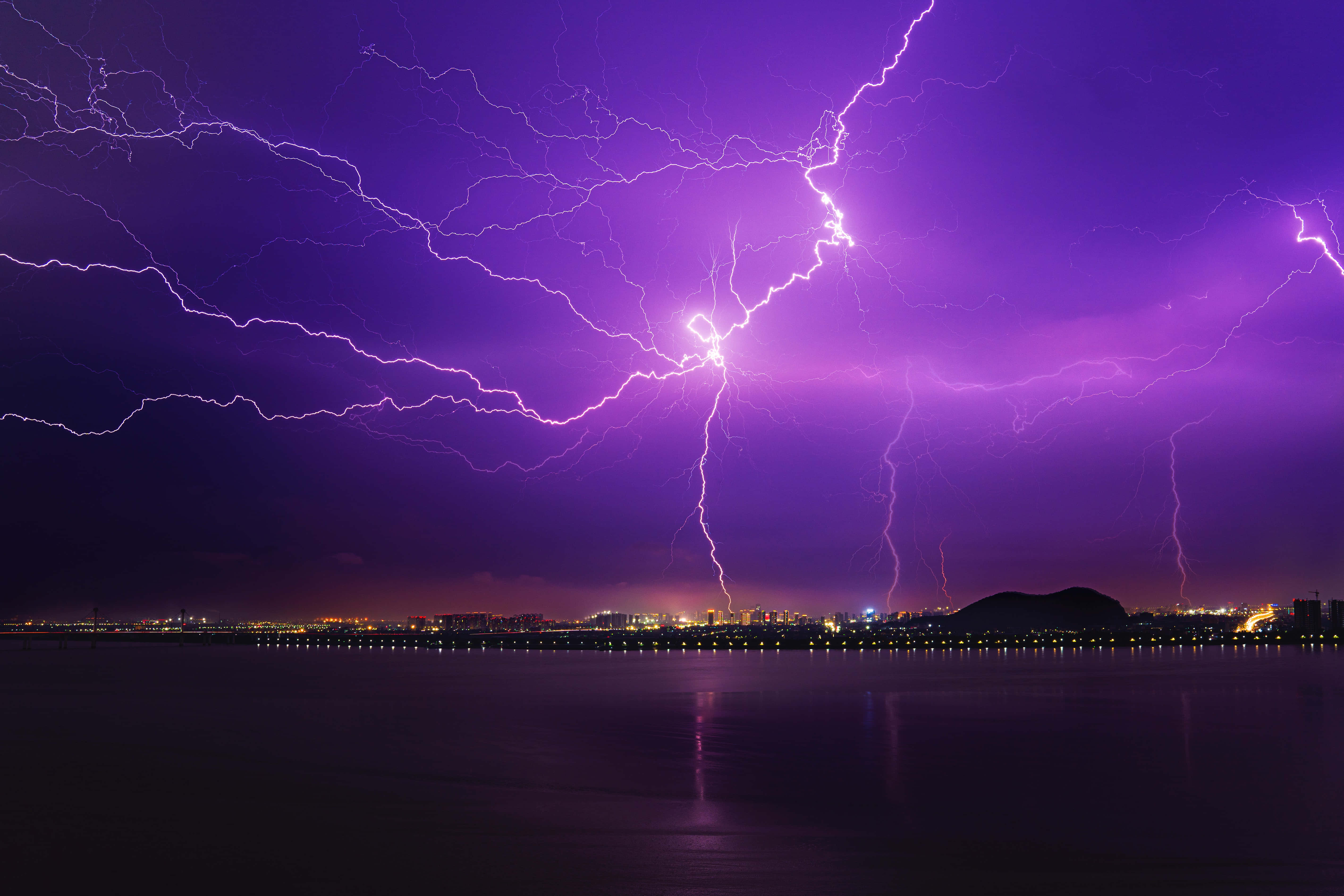 Purple Lightning 桌布 壁紙 — 鮮豔的紫色閃電在暴風雨的夜空中，於城市景觀和寧靜的水面上方叉開。