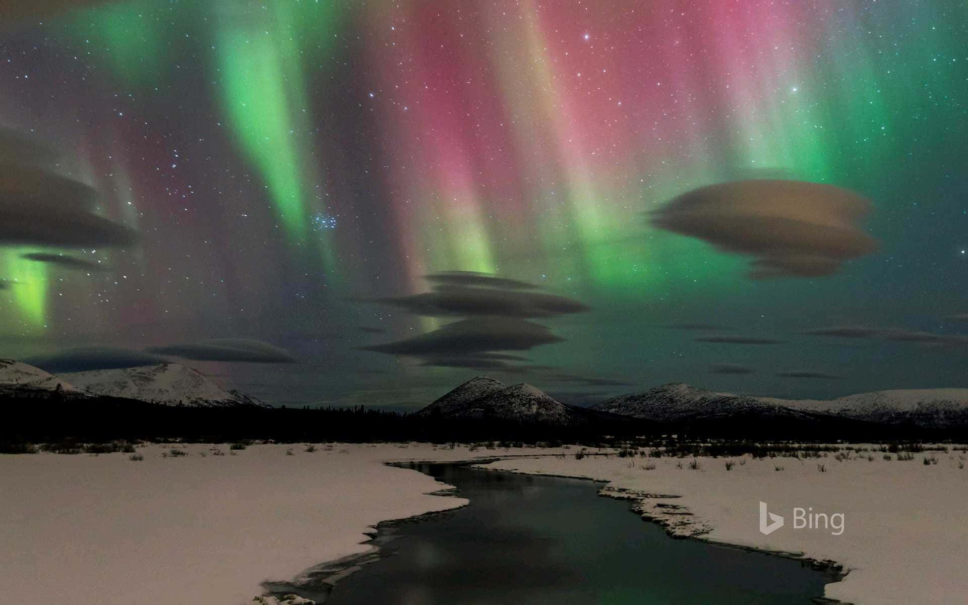 必應 桌布 壁紙 — 鮮豔的綠色和粉紅色極光在白雪覆蓋的山脈和冰凍的河流上方，在繁星點點的夜空下舞動。