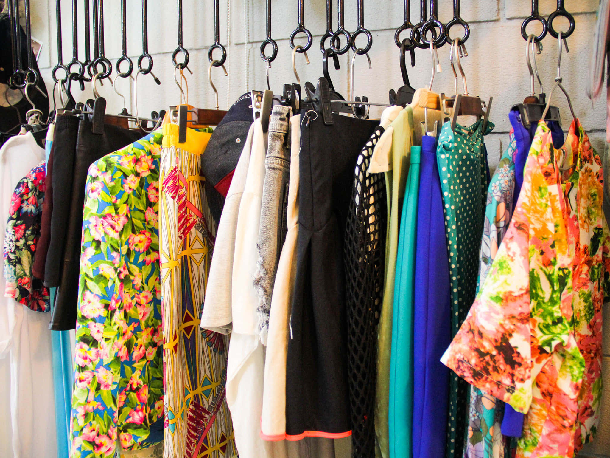 Clothes 桌布 壁紙 — 鮮豔的花卉和圖案衣物密集地掛在黑色掛鉤上，背景是精品店內有紋理的牆壁。