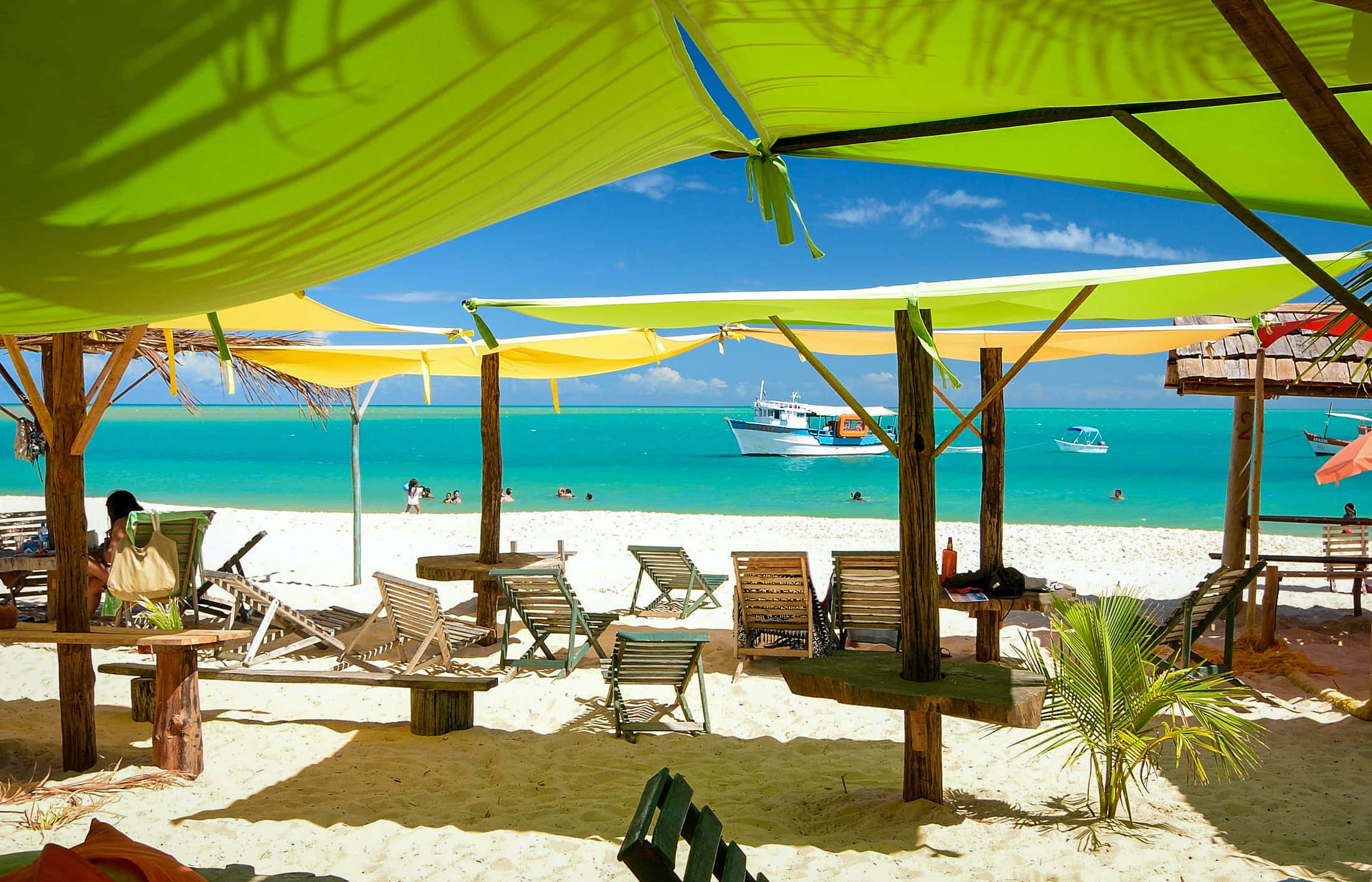 Beautiful Beach 桌布 壁紙 — 鮮豔的萊姆綠和黃色遮陽篷陰影覆蓋在木製躺椅上，位於純淨的白色沙灘上，背景是碧綠的海水和船隻，天空清澈湛藍。