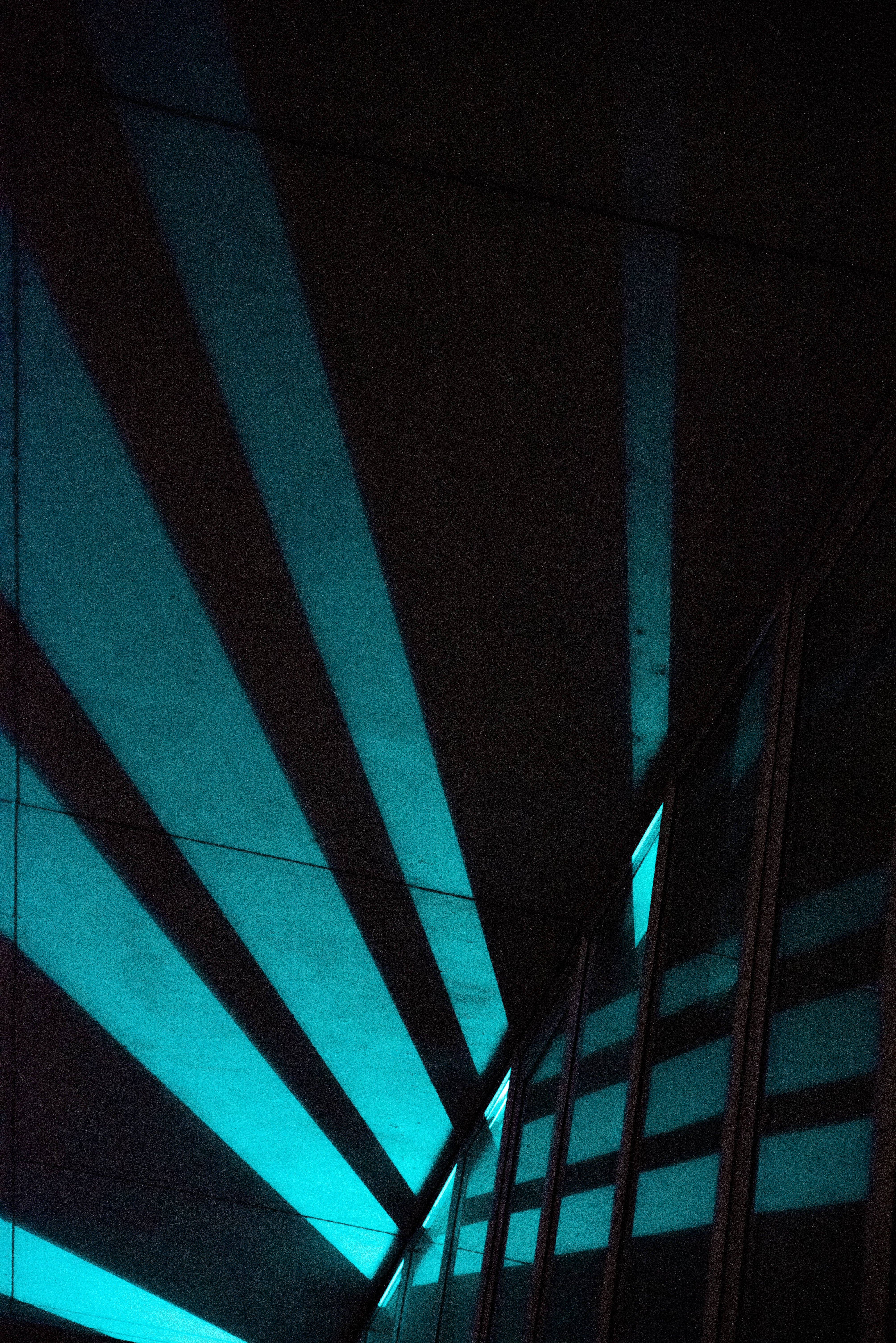 dark neon iPhone 桌布 壁紙 — 鮮豔的藍綠色光束劃破黑暗，在現代建築室內營造出戲劇性的幾何陰影。