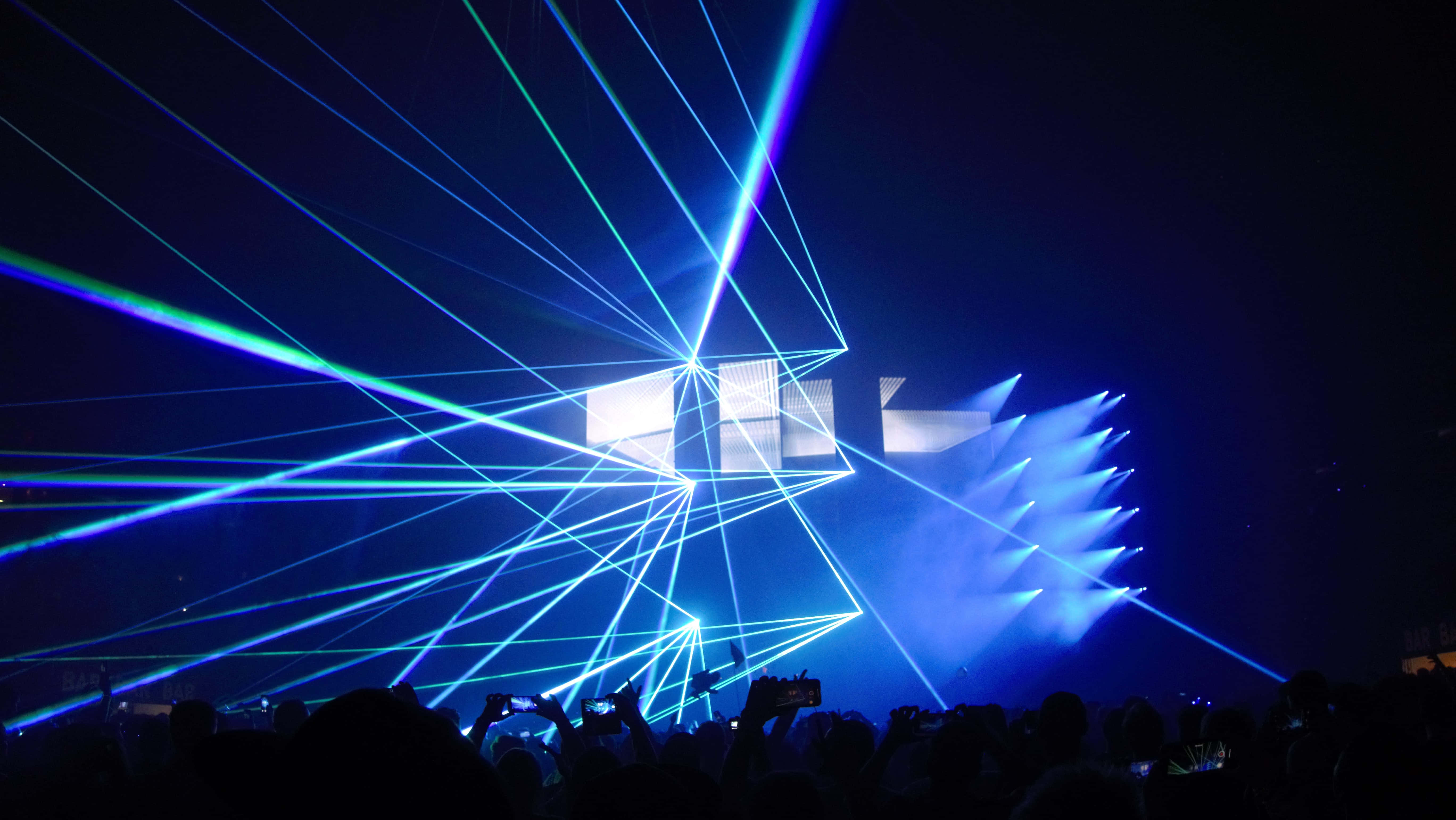 Laser 桌布 壁紙 — 鮮豔的藍色和綠色雷射光束在黑暗的演唱會舞台上交錯，剪影般的觀眾舉起手機捕捉這一壯觀景象。