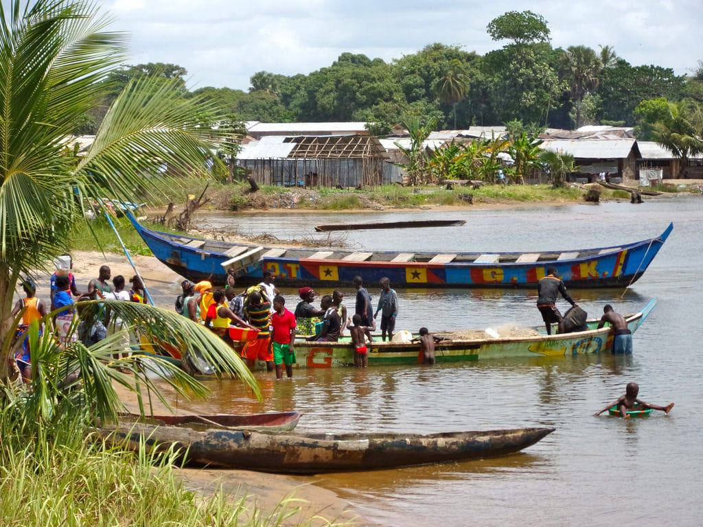 Liberia 桌布 壁紙 — 鮮豔的藍色和黃色船隻停泊在泥濘的河岸，人們在多雲的天空下裝載貨物。