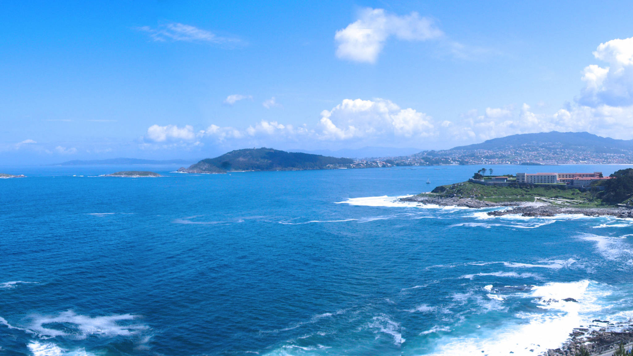 View 桌布 壁紙 — 鮮豔的藍色海浪拍打岩石海岸，天空明亮，雲朵散佈，靠近一個沿海小鎮。