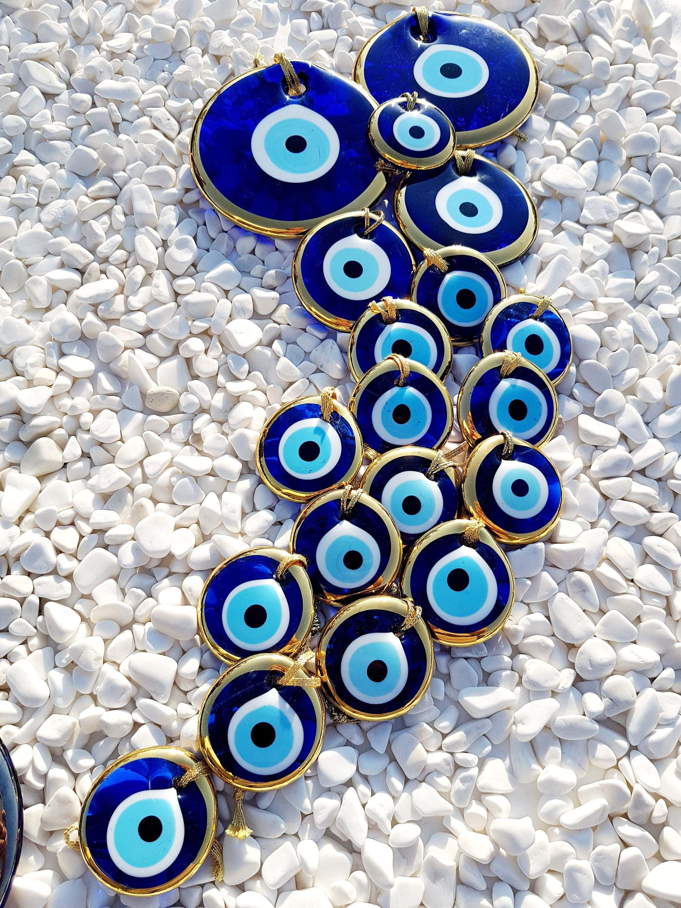 Evil Eye 桌布 壁紙 — 鮮豔的藍色邪眼護身符，帶有金色邊框，在明亮的陽光下以瀑布狀排列在白色鵝卵石上。