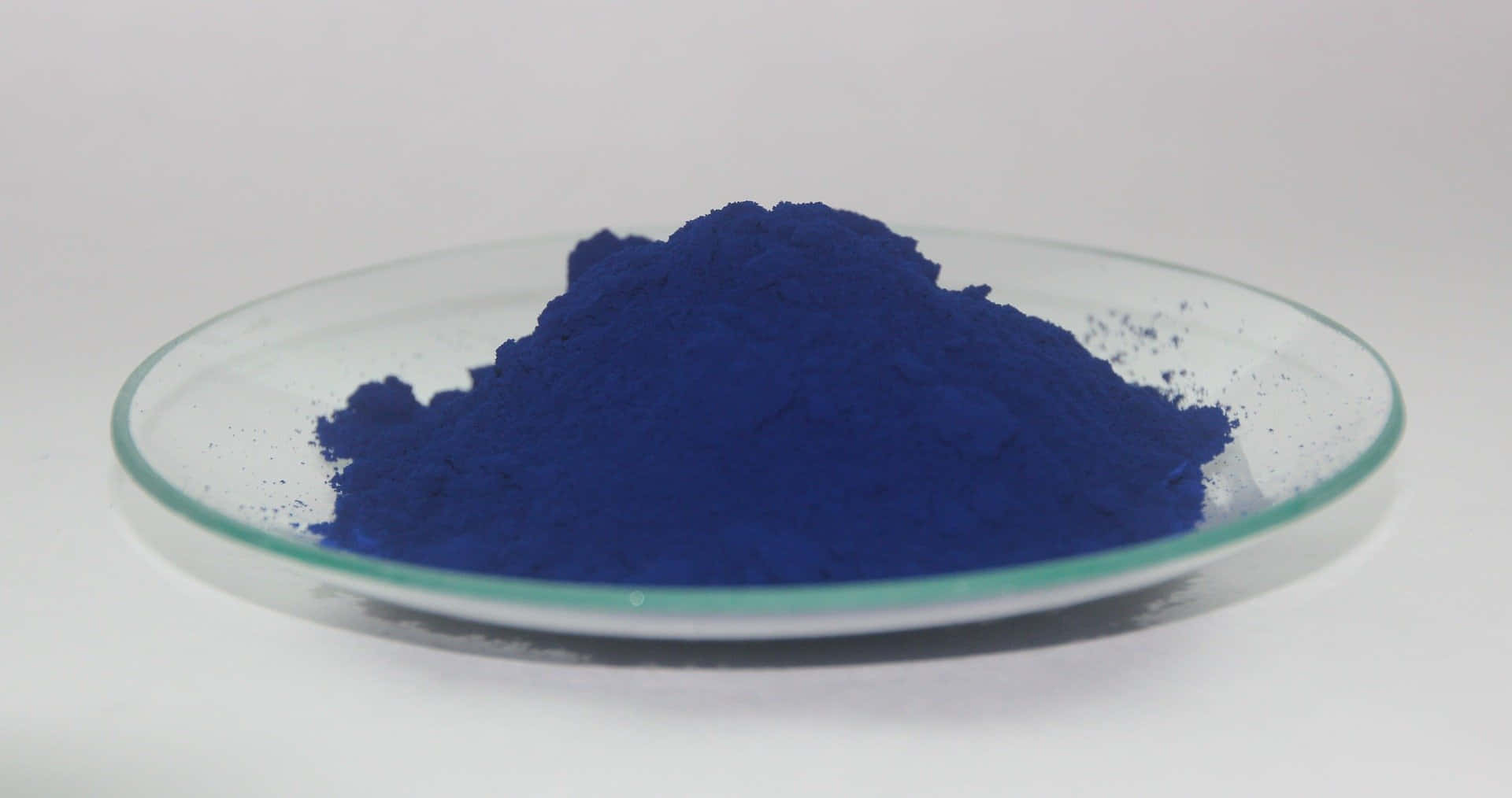 Indigo Blue 桌布 壁紙 — 鮮豔的鈷藍色粉末高高堆疊在透明玻璃培養皿中，背景為純白色。