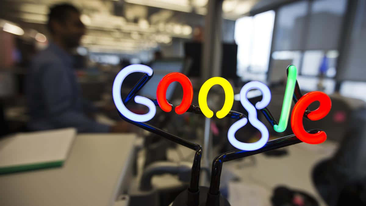 google 桌布 壁紙 — 鮮豔的霓虹Google標誌在前景發光，背景有模糊的辦公室員工。