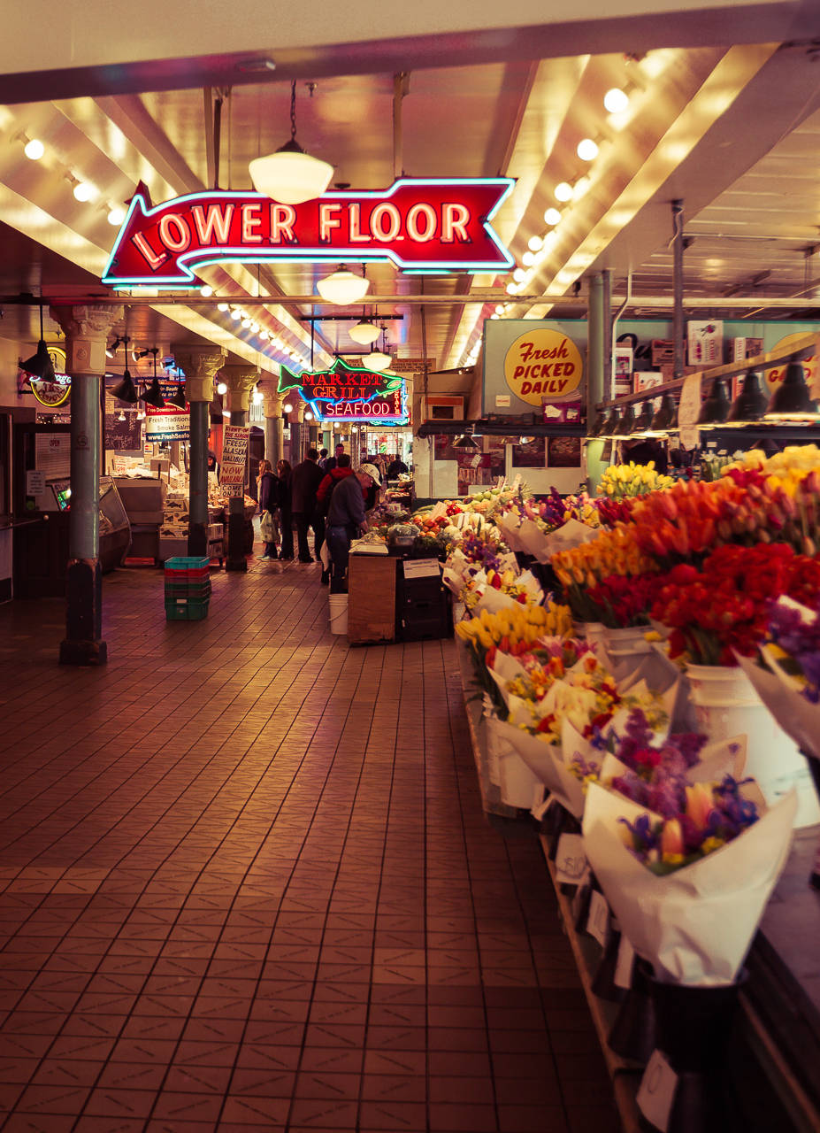 Pike Place Market 桌布 壁紙 — 鮮豔的霓虹燈照亮了一條繁忙的室內市場通道，兩旁擺滿了色彩繽紛的花束和購物者。
