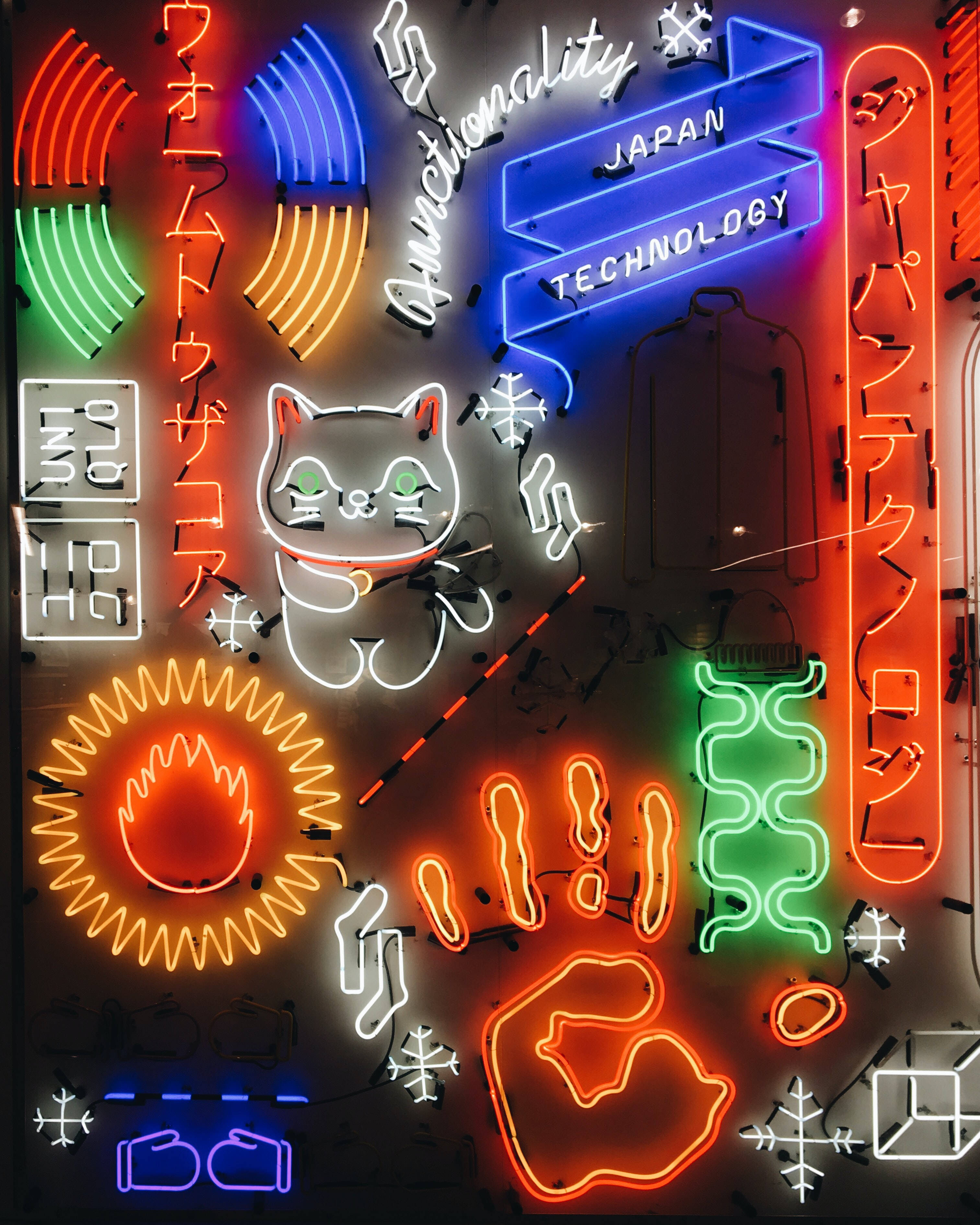 Japanese Aesthetic 桌布 壁紙 — 鮮豔的霓虹燈牌以日文和英文顯示，在黑暗的牆壁上發出紅、藍、綠、橙色的光芒，帶有貓和科技主題。