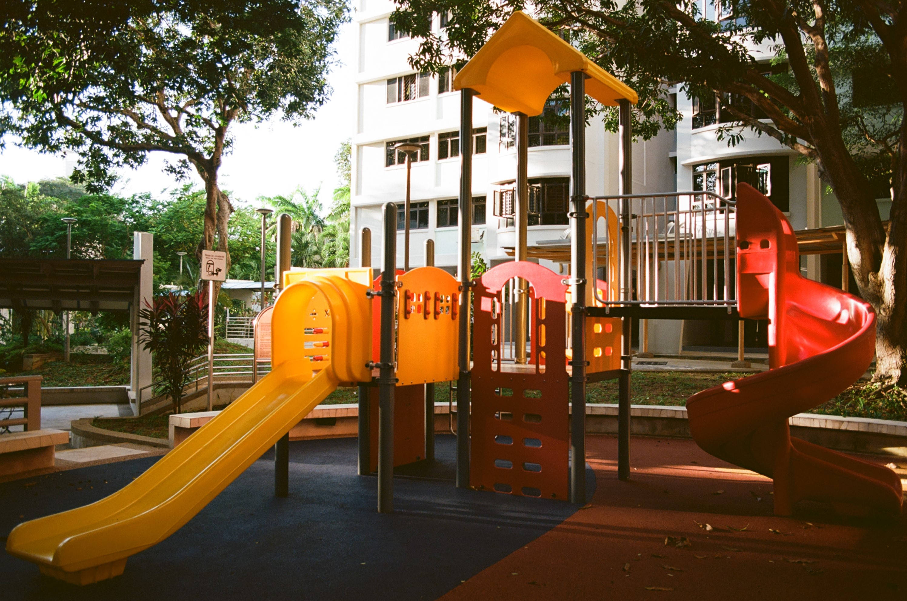 Playground 桌布 壁紙 — 鮮豔的黃色和紅色遊樂場滑梯在溫暖的陽光下閃耀，位於樹蔭遮蔽的市區公園。