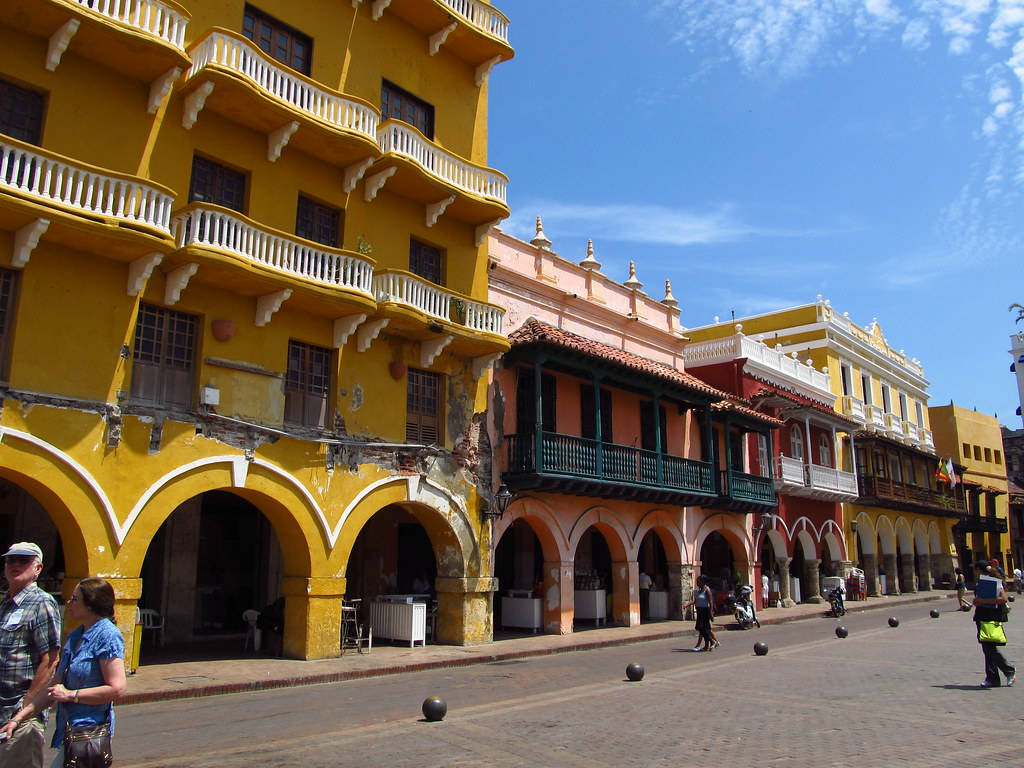 Colombia 桌布 壁紙 — 鮮黃色和粉紅色的殖民風格建築物搭配白色陽台，排列在一條陽光普照的鵝卵石街道上，上方是清澈的藍天。