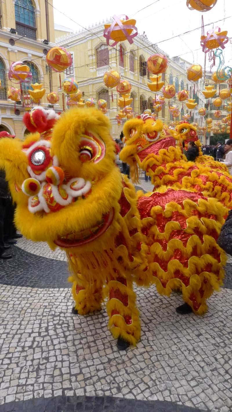 春節 桌布 壁紙 — 鮮黃色和紅色的舞獅服裝在懸掛燈籠下的鵝卵石街道上表演。
