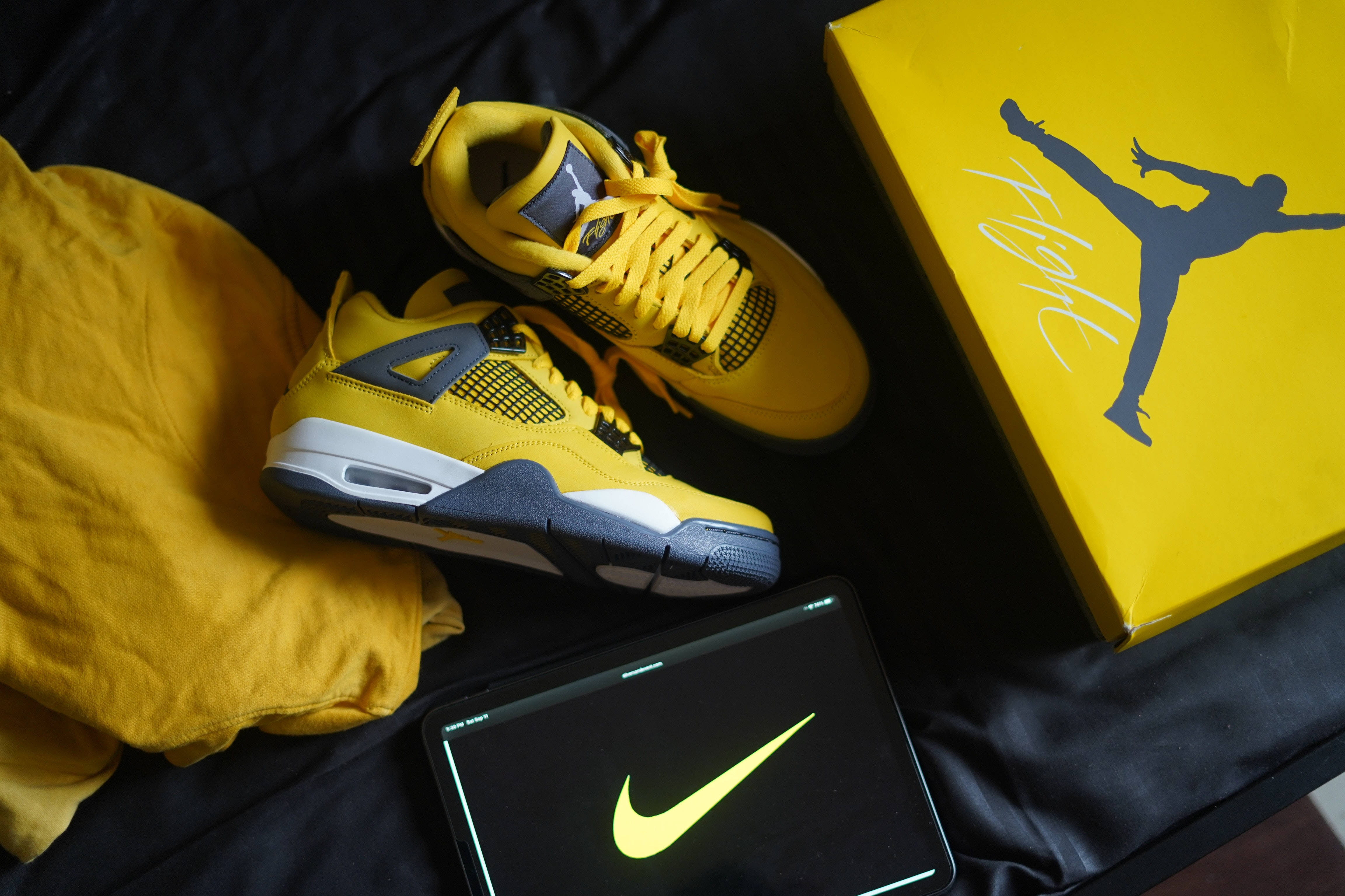 Nike Shoes 桌布 壁紙 — 鮮黃色的Jordan 4運動鞋放在盒子旁邊，旁邊的平板電腦上顯示發光的Nike標誌，置於深色布料上。