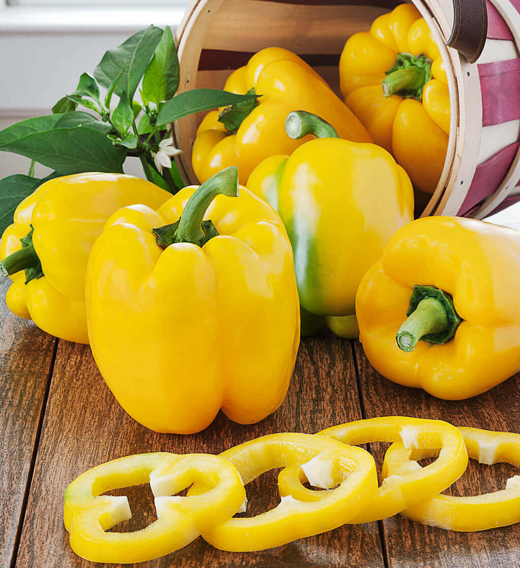 Yellow Bell Pepper 桌布 壁紙 — 鮮黃色的甜椒（完整和切片）從木籃中溢出，落在一個帶有新鮮綠葉的鄉村風格木桌上。