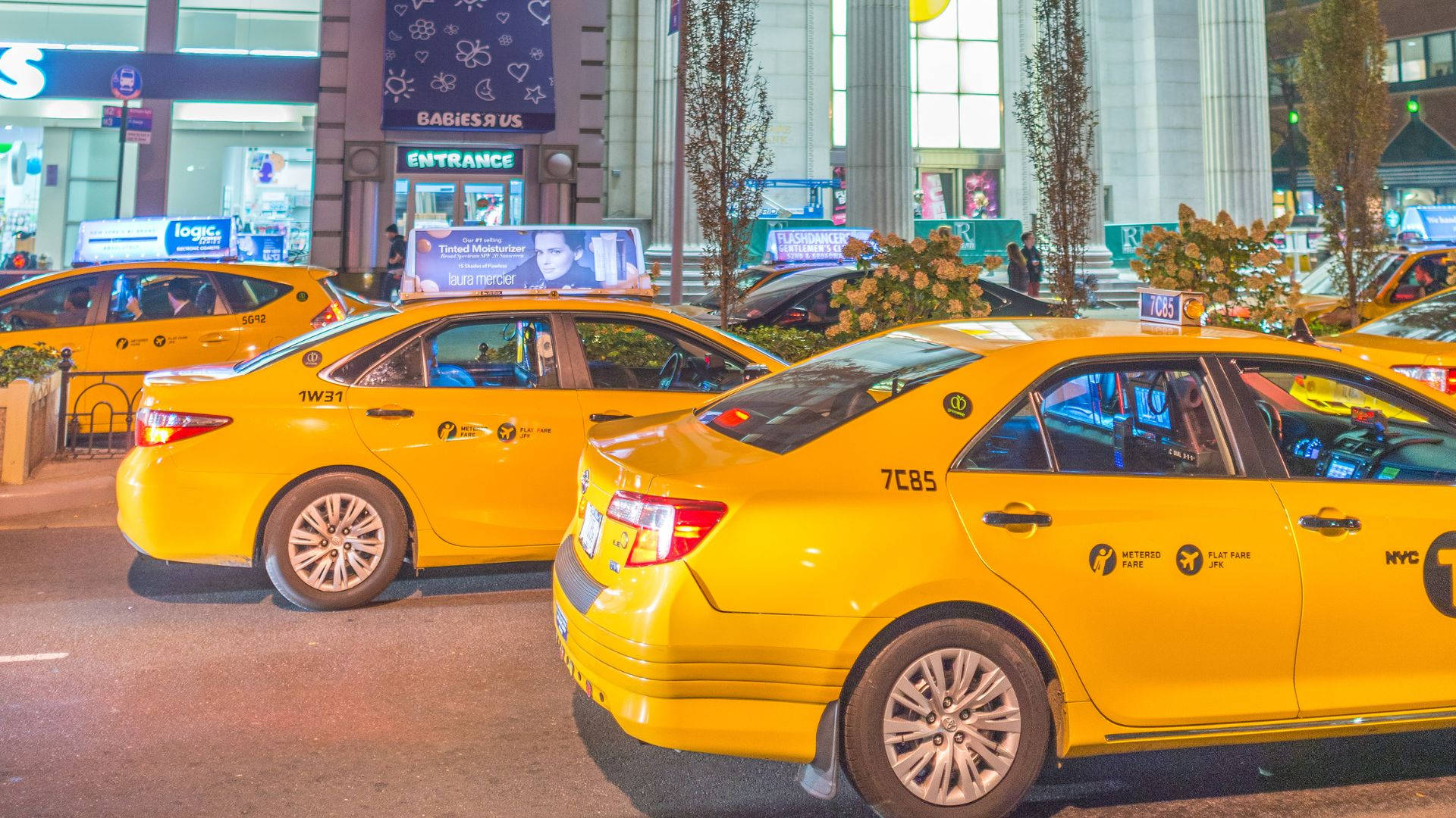 taxi 桌布 壁紙 — 鮮黃色的紐約計程車在夜晚排列在繁忙的城市街道上，伴隨著發光的商店櫥窗和行人。