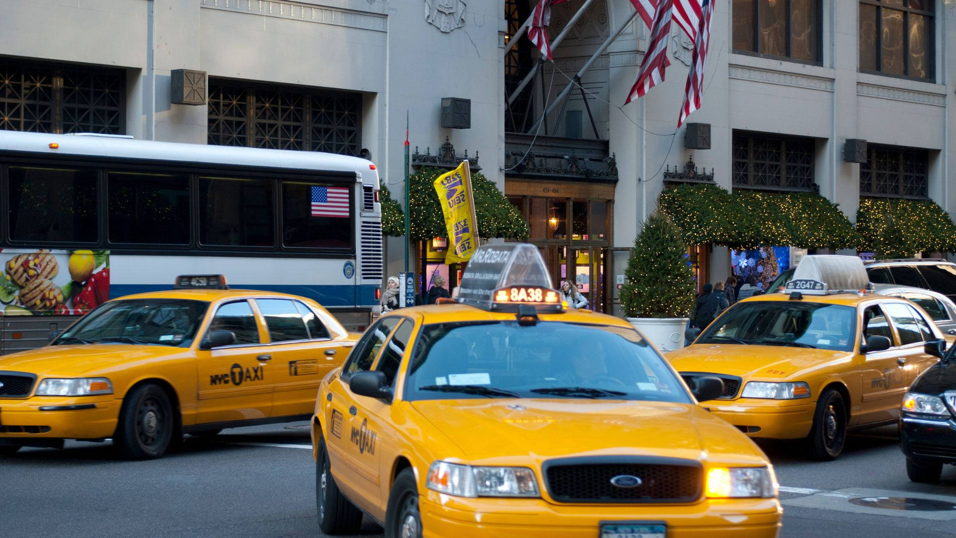 taxi 桌布 壁紙 — 鮮黃色的紐約計程車在日光下穿梭於繁忙的城市街道，經過節日綠化和歷史建築。