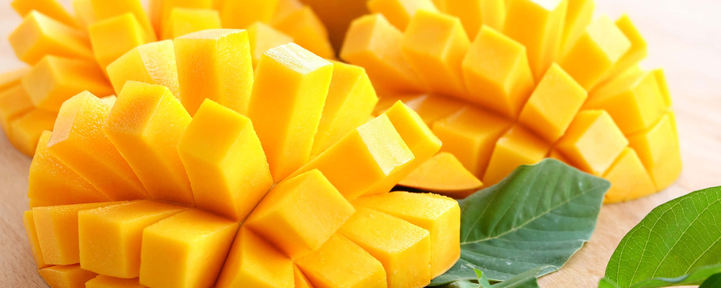 Mango 桌布 壁紙 — 鮮黃色芒果肉切成整齊的方塊，以扇形排列在木質表面上，旁邊放上新鮮的綠葉。