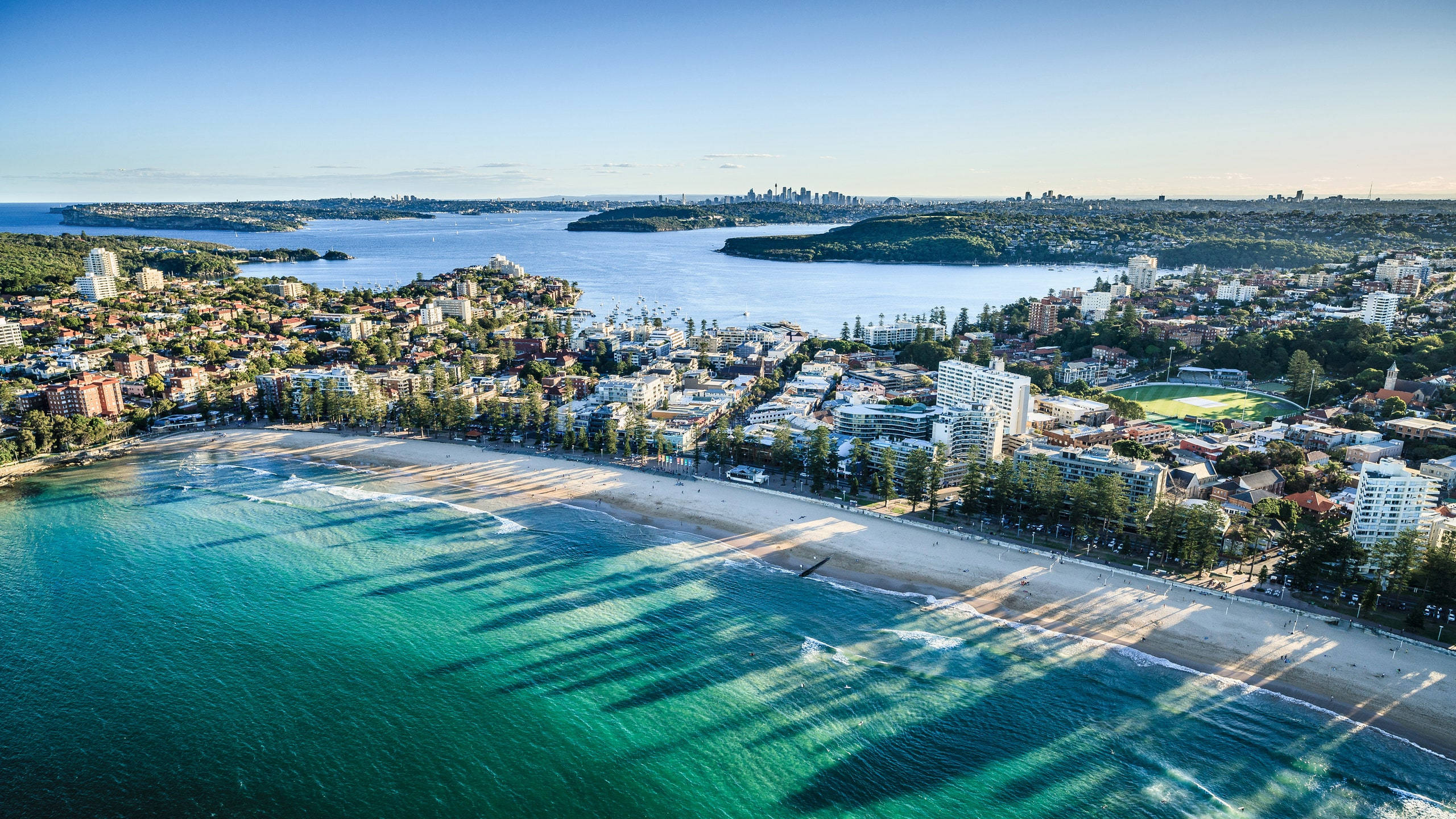 Sydney 桌布 壁紙 — 鳥瞰一個充滿活力的沿海城市，碧綠的海水在沙灘上投下長長的影子，沙灘兩旁是建築物和綠化，天空清澈湛藍。
