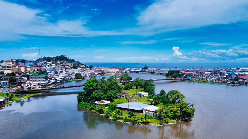 Liberia 桌布 壁紙 — 鳥瞰一個充滿活力的沿海小鎮，被一條寬闊的河流分隔，中央有一個鬱鬱蔥蔥的綠色島嶼，在明亮的藍天下。
