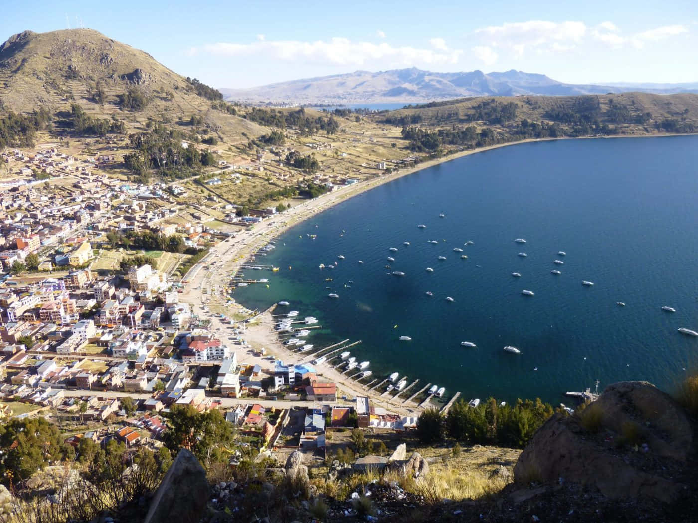 Bolivia 桌布 壁紙 — 鳥瞰一個充滿活力的湖邊小鎮，依偎在起伏的丘陵之間，深藍色的水面上點綴著船隻，天空明亮。