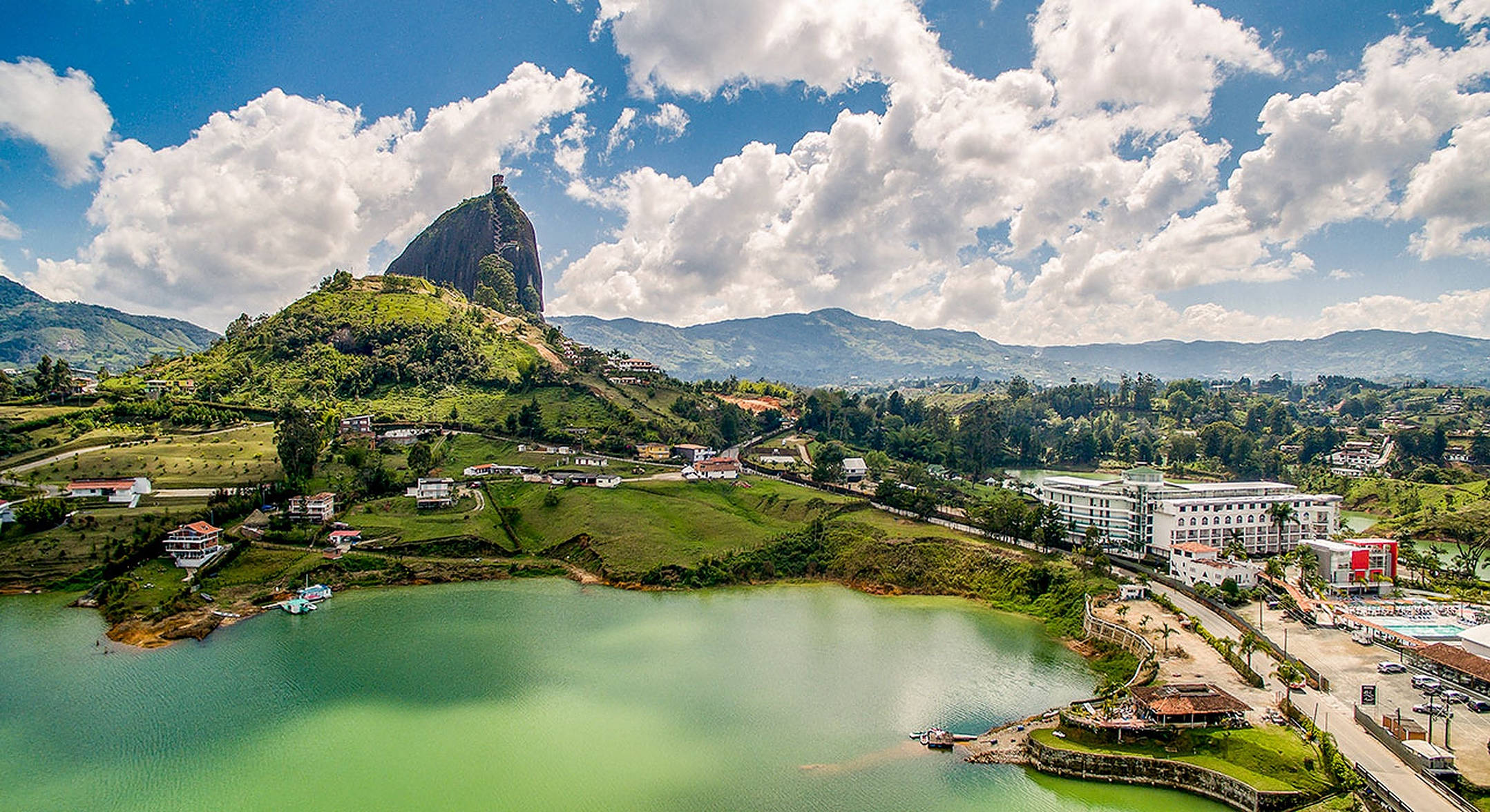 Colombia 桌布 壁紙 — 鳥瞰一個充滿活力的綠色湖泊，旁邊有一座巨大的岩石山丘，在明亮的藍天和蓬鬆的白雲下。