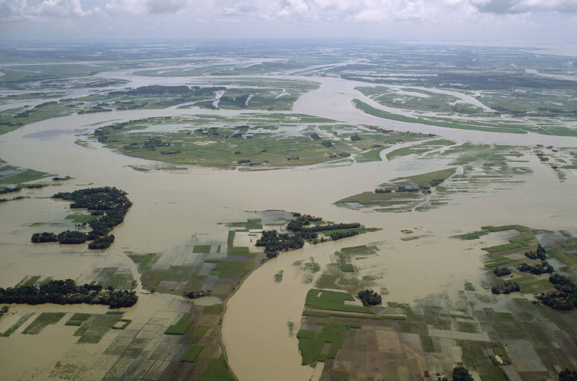 river 桌布 壁紙 — 鳥瞰一個廣闊的洪水氾濫河流三角洲，泥濘的水道蜿蜒穿過拼布般的綠色田野和散落的樹木，天空多雲。