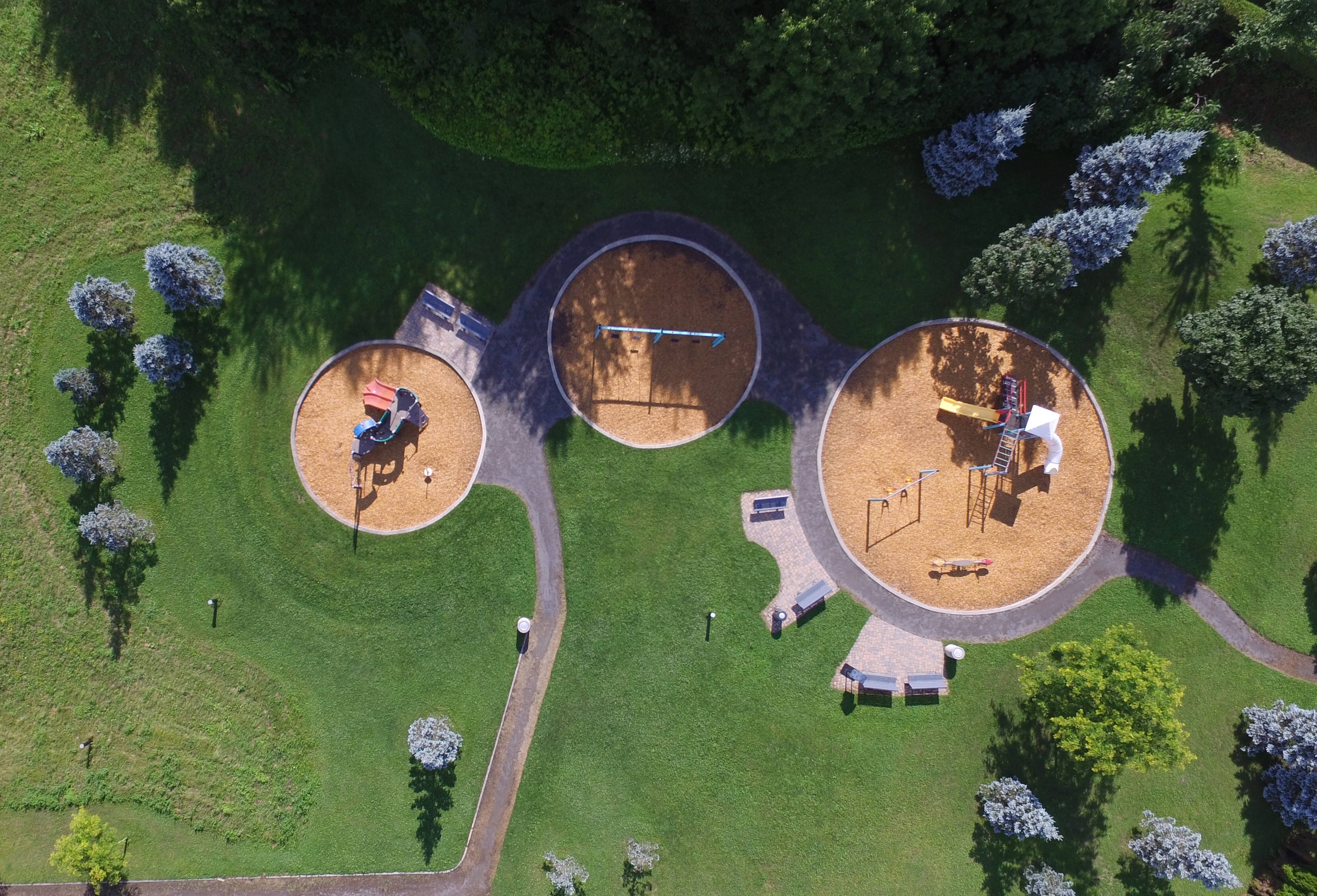 Playground 桌布 壁紙 — 鳥瞰一個現代遊樂場，有三個圓形沙坑，周圍環繞著茂密的綠草和樹木，在明亮的陽光下。