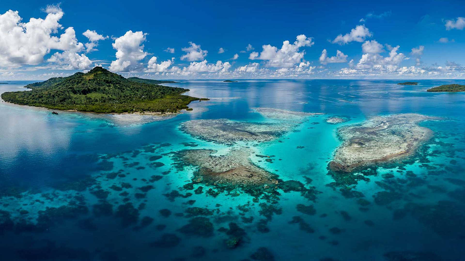 Micronesia 桌布 壁紙 — 鳥瞰一個鬱鬱蔥蔥的綠色島嶼，周圍環繞著充滿活力的綠松石珊瑚礁，在明亮的藍天下有蓬鬆的白雲。