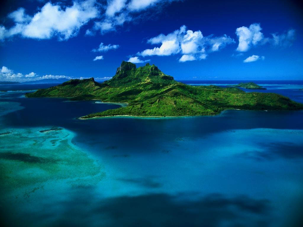 海洋 桌布 壁紙 — 鳥瞰一個鬱鬱蔥蔥的綠色火山島，周圍環繞著充滿活力的碧綠和深藍色海水，天空明亮，雲朵散落。