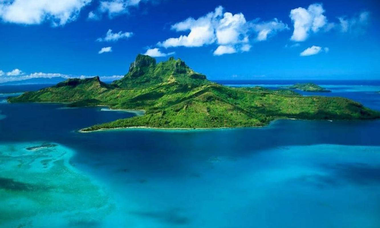 Haiti 桌布 壁紙 — 鳥瞰一個鬱鬱蔥蔥的綠色火山島，周圍環繞著充滿活力的碧綠海水，上方是明亮的藍天和蓬鬆的白雲。