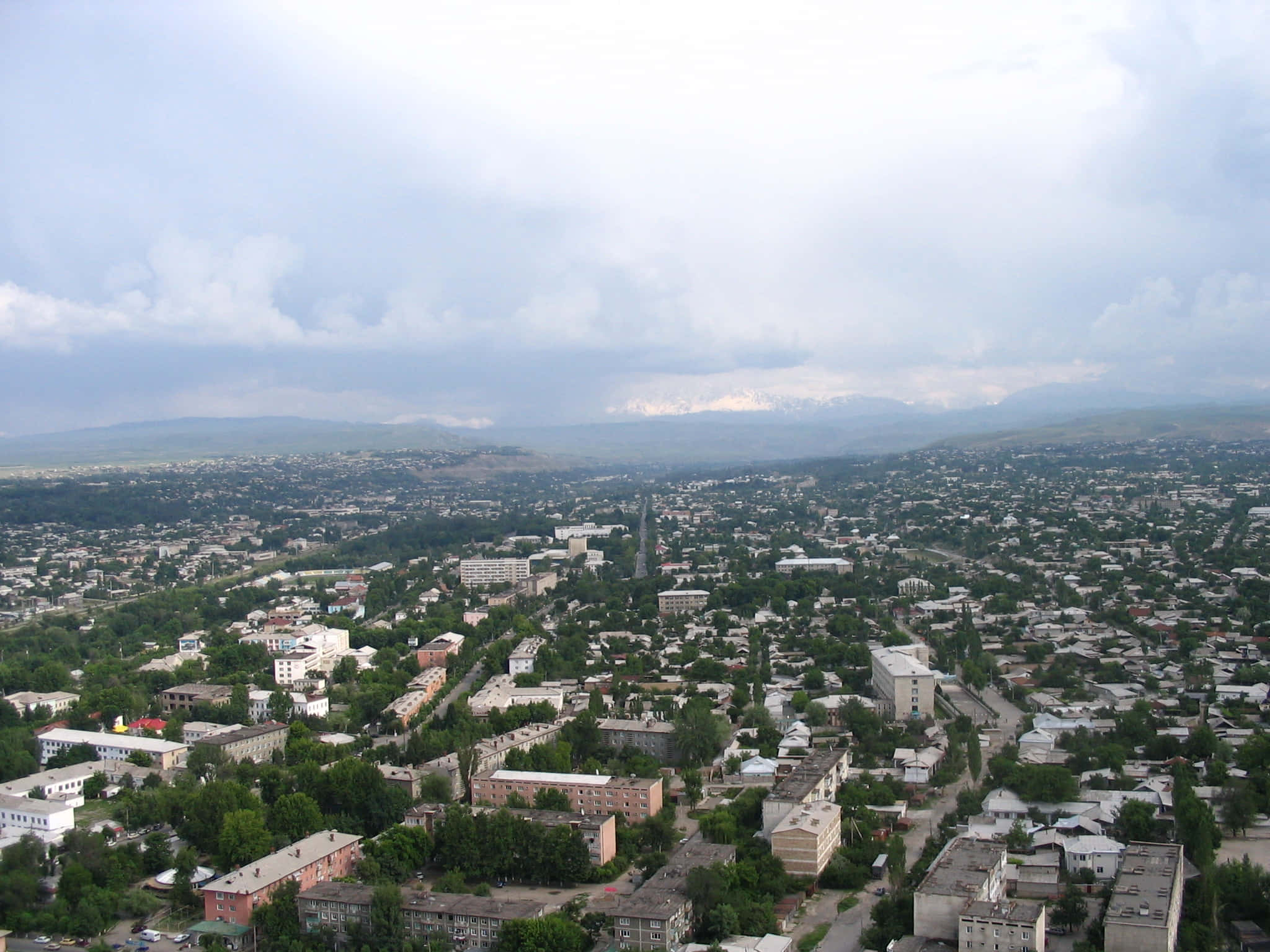 Kyrgyzstan 桌布 壁紙 — 鳥瞰一座廣闊的城市，坐落在綠丘之間，積雪的山峰在陰天天空下清晰可見。