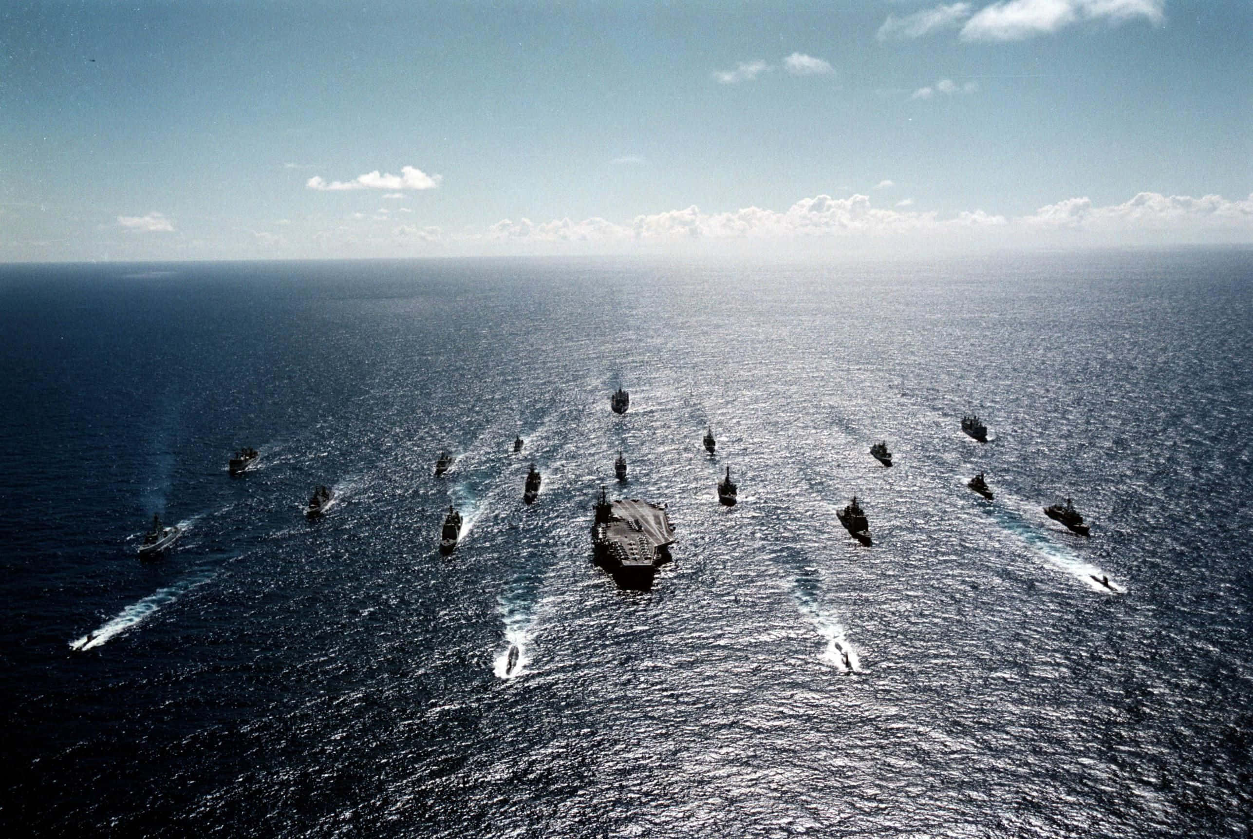 Navy 桌布 壁紙 — 鳥瞰一支以航空母艦為中心的海軍艦隊，在晴朗天空下的廣闊藍色海洋上編隊航行。
